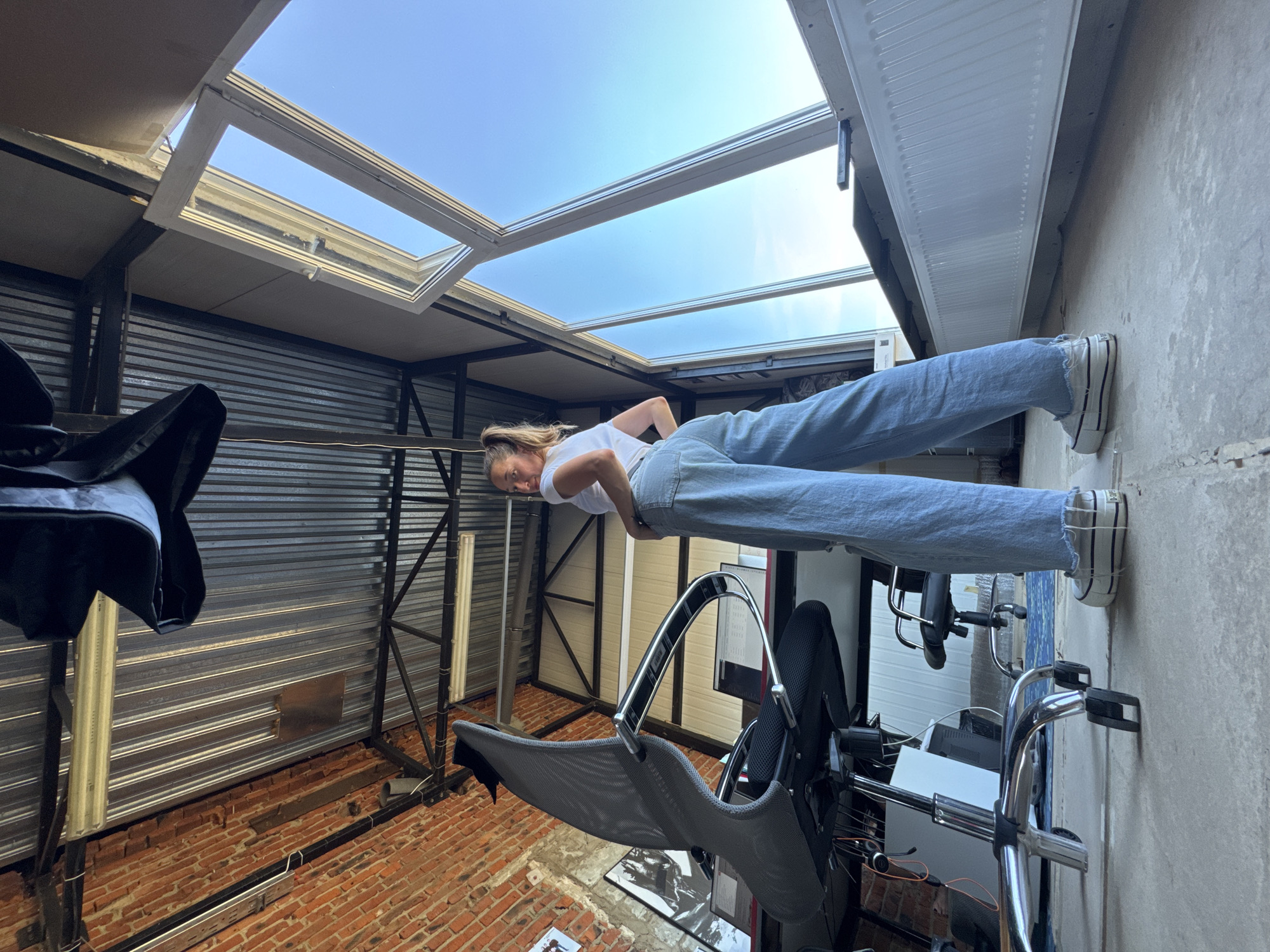 A woman stands in a room with a skylight. She wears jeans and a white shirt, and her posture is slightly tilted to her left. The room appears to be a garage or warehouse-like space. The walls are constructed from corrugated metal sheets, with brickwork visible on the lower sections. A metal framework supports lighting fixtures against the metal wall. The floor is brick. Modern office furniture including a chair and computer are also in the room. The skylight lets in a view of the sky. Without additional information or data points, I am unable to establish the location as Chaikovsky, Russia.