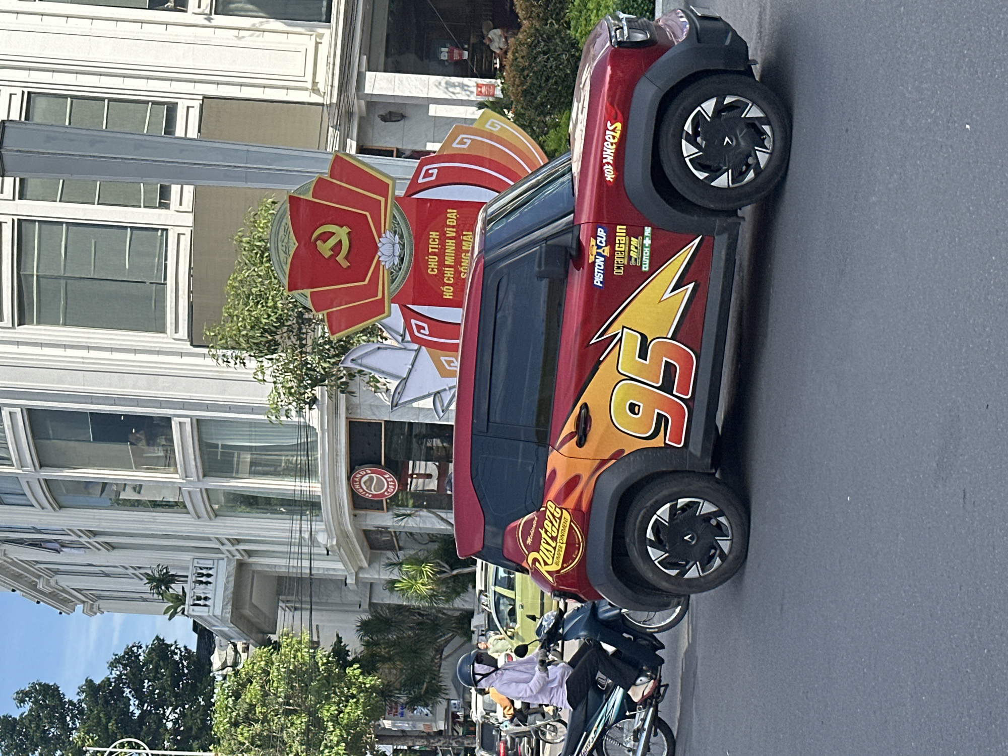 An eye-level shot captures a vibrant, customized red car resembling Lightning McQueen from the "Cars" movie, parked on a sunlit urban street in what appears to be Vietnam. The car is adorned with its iconic "95" number, yellow lightning bolt graphics, and fictional sponsor decals such as "Rusteze Bumper Ointment," "Piston Cup," "Octane Gain," "RPM," and "Clutch-Aid." A "Hot Wheels" logo is also visible near the front wheel.
Adjacent to the car stands a prominent red and gold decorative structure, featuring the hammer and sickle communist emblem and Vietnamese text that reads: "CHỦ TỊCH HỒ CHÍ MINH VĨ ĐẠI SỐNG MÃI" (Chairman Ho Chi Minh, the Great, Lives Forever). This structure signifies a political commemoration or celebration.
The street scene is active, with several motorbikes and their riders visible, one person in the foreground wearing a helmet and a white shirt. In the background, multi-story buildings with balconies and intricate architectural details line the street. A recognizable "HIGHLANDS COFFEE" sign is visible on one building, further indicating the Vietnamese setting. Lush green trees are scattered along the pavement. The bright lighting suggests it is daytime with clear weather.