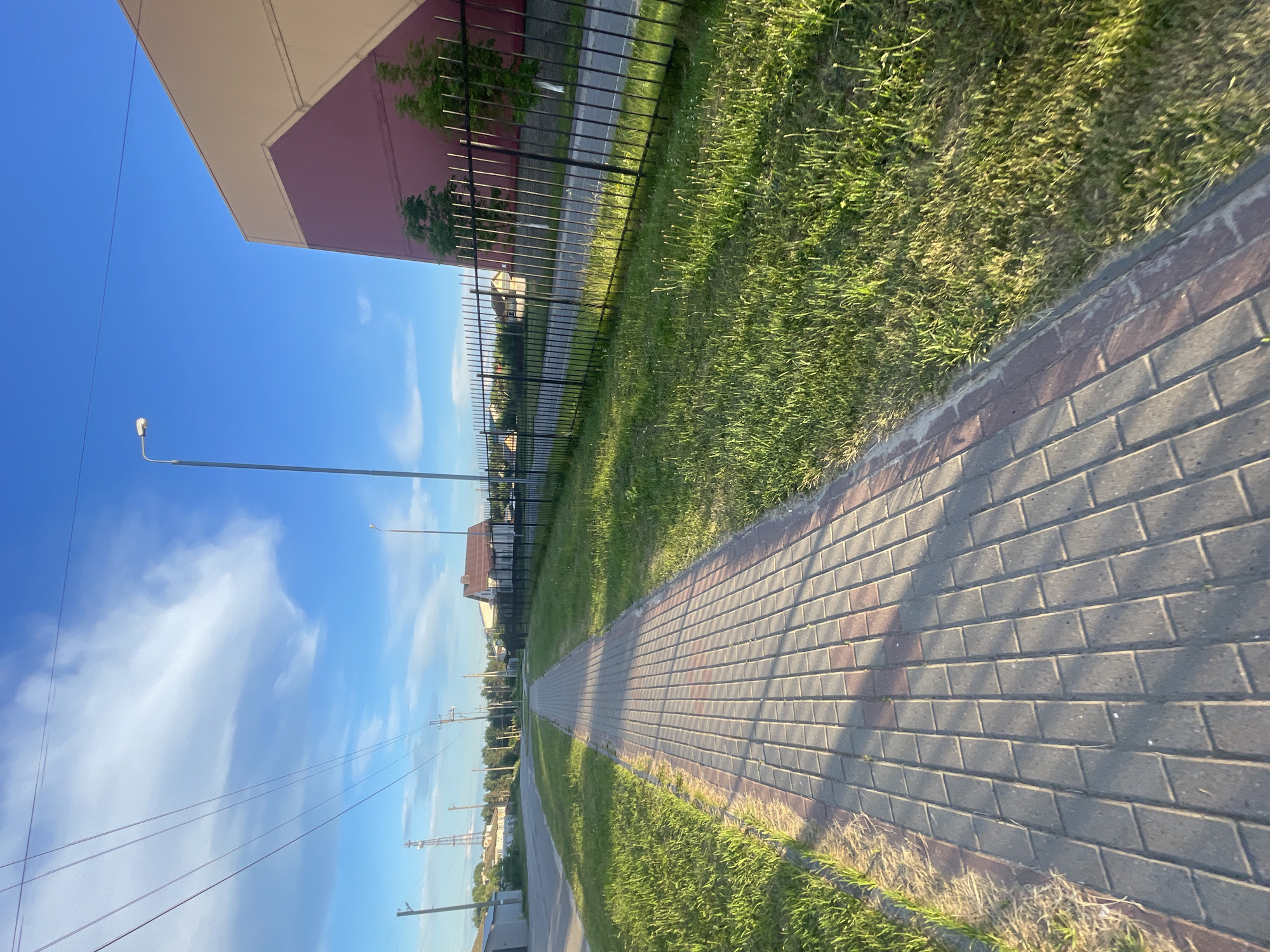 This image captures a paved pedestrian walkway in Krasnye Baki, Russia, on a bright, sunny day. The walkway, constructed of gray and reddish-brown interlocking pavers, runs diagonally from the lower left to the upper right of the frame, bordered by a strip of dry grass and then a well-maintained green lawn. Beyond the lawn, a black metal fence with vertical bars separates the walkway area from a road and a cluster of residential buildings with red-tiled roofs. To the right, a large building with a red and beige facade stands, with two young trees in front of it. In the background, utility poles with wires are visible, as well as a cell tower in the distance. The sky is a vibrant blue with scattered white clouds. There are no people or animals in the image.