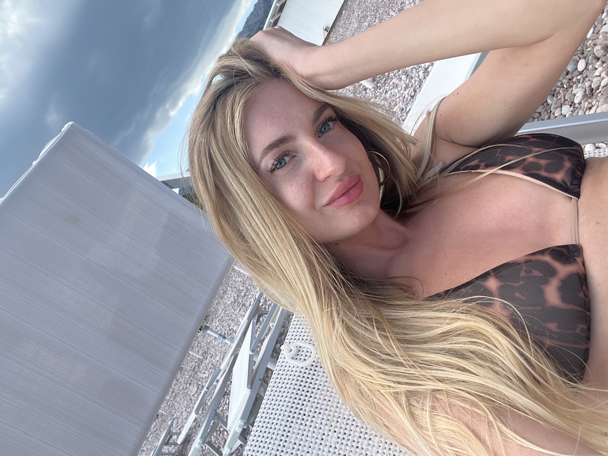 A woman with long, blonde hair lies on a white lounge chair. She is wearing a brown animal-print bikini top. Her right arm is raised, with her hand touching her hair. She looks directly at the camera with blue eyes. Gold hoop earrings are visible. The lounge chair is on a pebbled surface. In the background is a cloudy sky and buildings with a mountain. The city is Sveti Stefan, Montenegro.