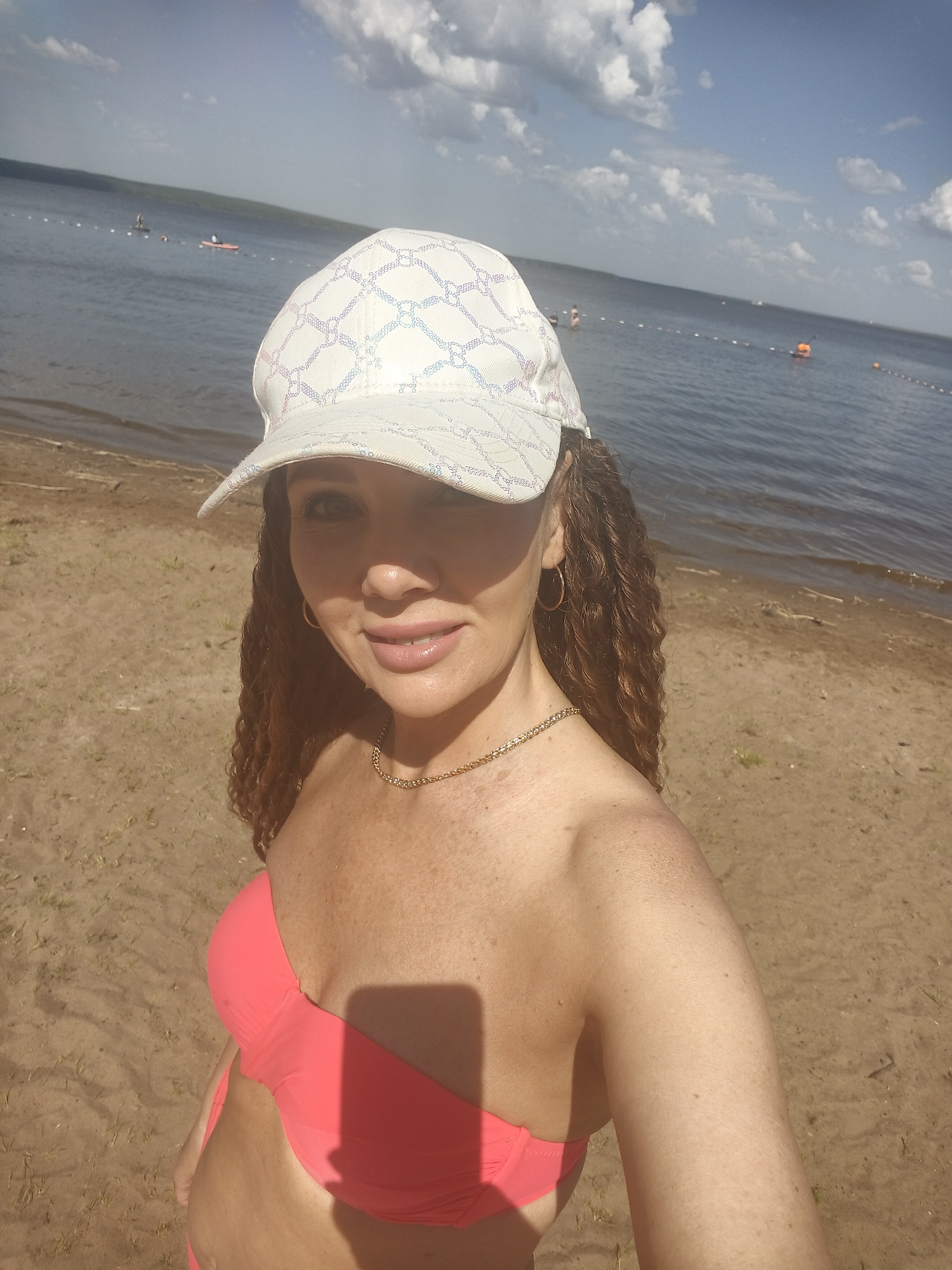 A woman stands on a sandy beach on a sunny day. She wears a white baseball cap, a pink bikini top, and a gold necklace and earrings. The beach stretches out behind her, leading to a body of water dotted with distant figures and buoys. The sky above is partly cloudy. The location is Unknown, Unknown.