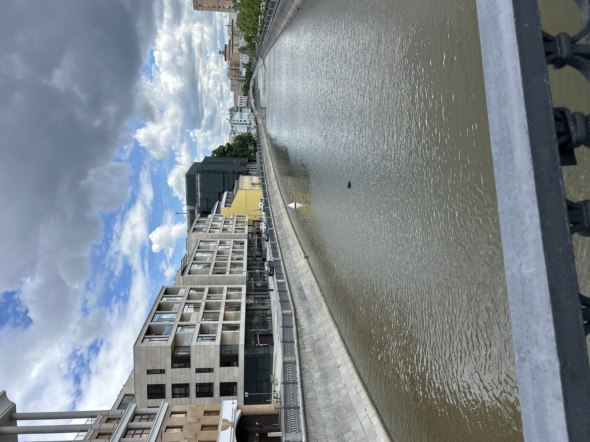 A photograph shows a body of water, likely a river or canal, flanked by urban infrastructure. The image has been rotated, with the sky and clouds occupying the upper left quadrant. Buildings line the left side of the waterway, exhibiting modern architectural designs with rectangular facades and numerous windows. The water's surface is textured with ripples, reflecting the ambient light. A small, dark object floats in the water. A stone embankment runs along the water's edge, separating it from the buildings and a street lined with parked vehicles. A metal railing is visible on the right side of the frame, suggesting the vantage point is from a bridge or elevated walkway. The sky is a mix of blue and heavy clouds. The overall scene presents an urban waterscape in daylight.