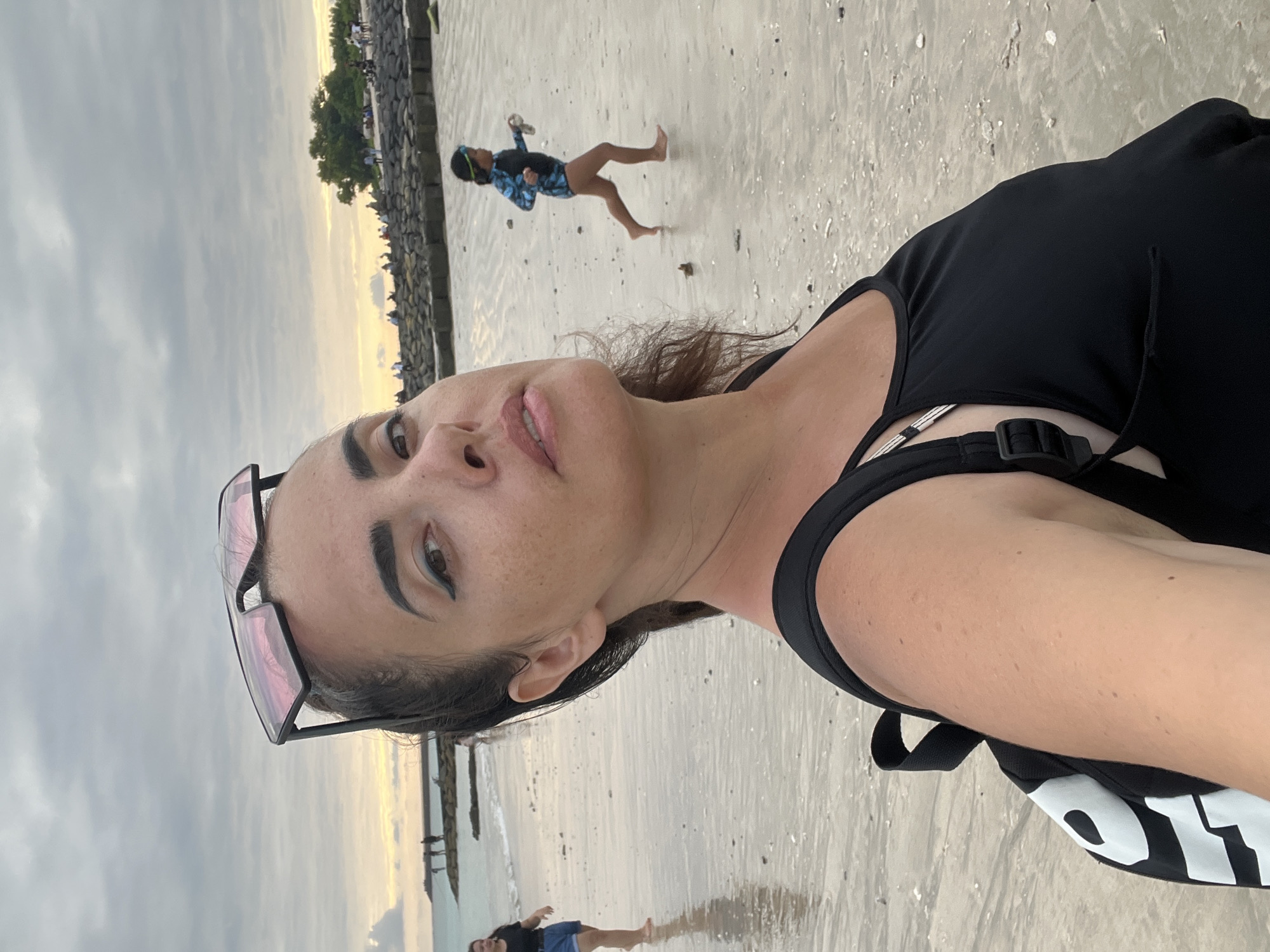 A woman is taking a selfie on a beach in Kuta, Indonesia, with a cloudy sky and gentle waves in the background. She is wearing a black tank top and a black backpack. Her sunglasses are perched on top of her head. In the background, a child in a blue and black swimsuit runs along the wet sand. Other people can be seen in the distance, some walking along the shore and others gathered near a rocky embankment. The beach is mostly wet sand with scattered shells and debris.