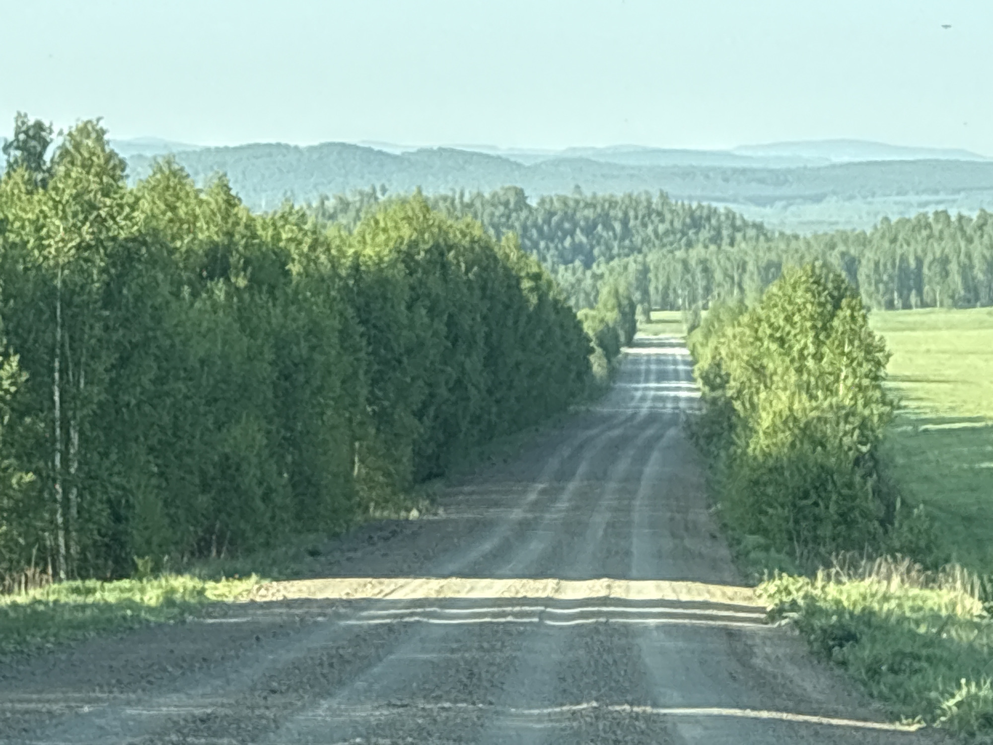 A long dirt road stretches into the distance, lined with green trees on both sides. In the background, layers of green hills and mountains fade into the hazy sky. Green grass borders the road on the right. The location is Unknown, Unknown.