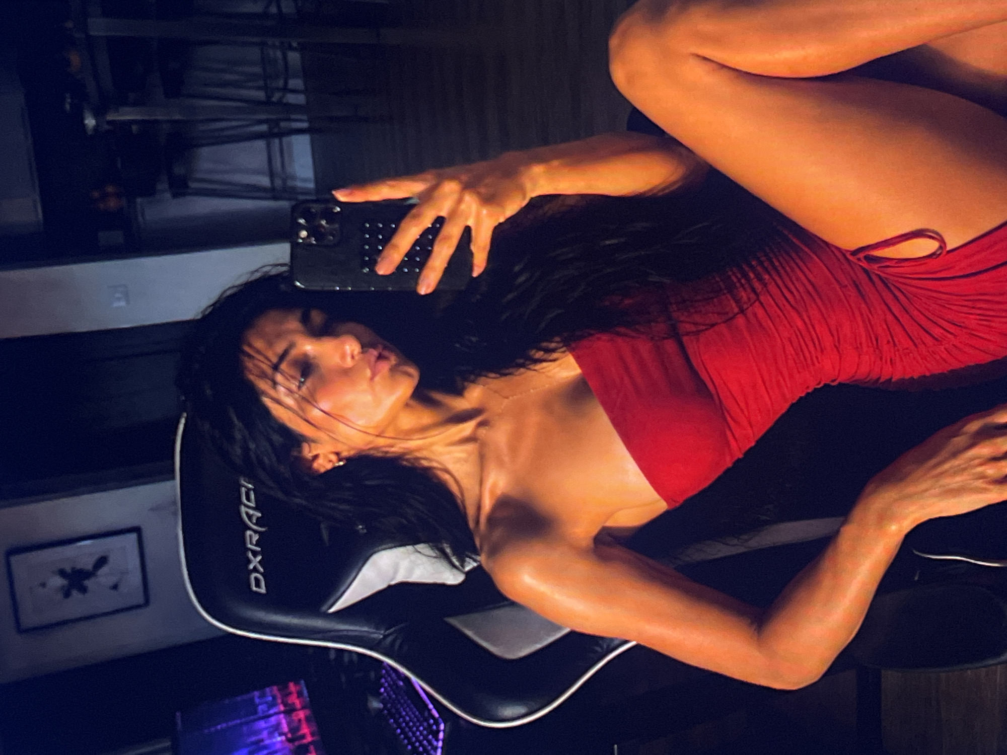 A woman with long, dark hair is seated in a black gaming chair, holding a smartphone in her right hand to take a mirror selfie. She is wearing a strapless red dress that is ruched on the side. Her left arm rests on her lap, and her legs are crossed. The background is dimly lit, with a computer monitor displaying a city skyline on the left and kitchen counter and stools on the right. A framed picture hangs on the wall behind her.