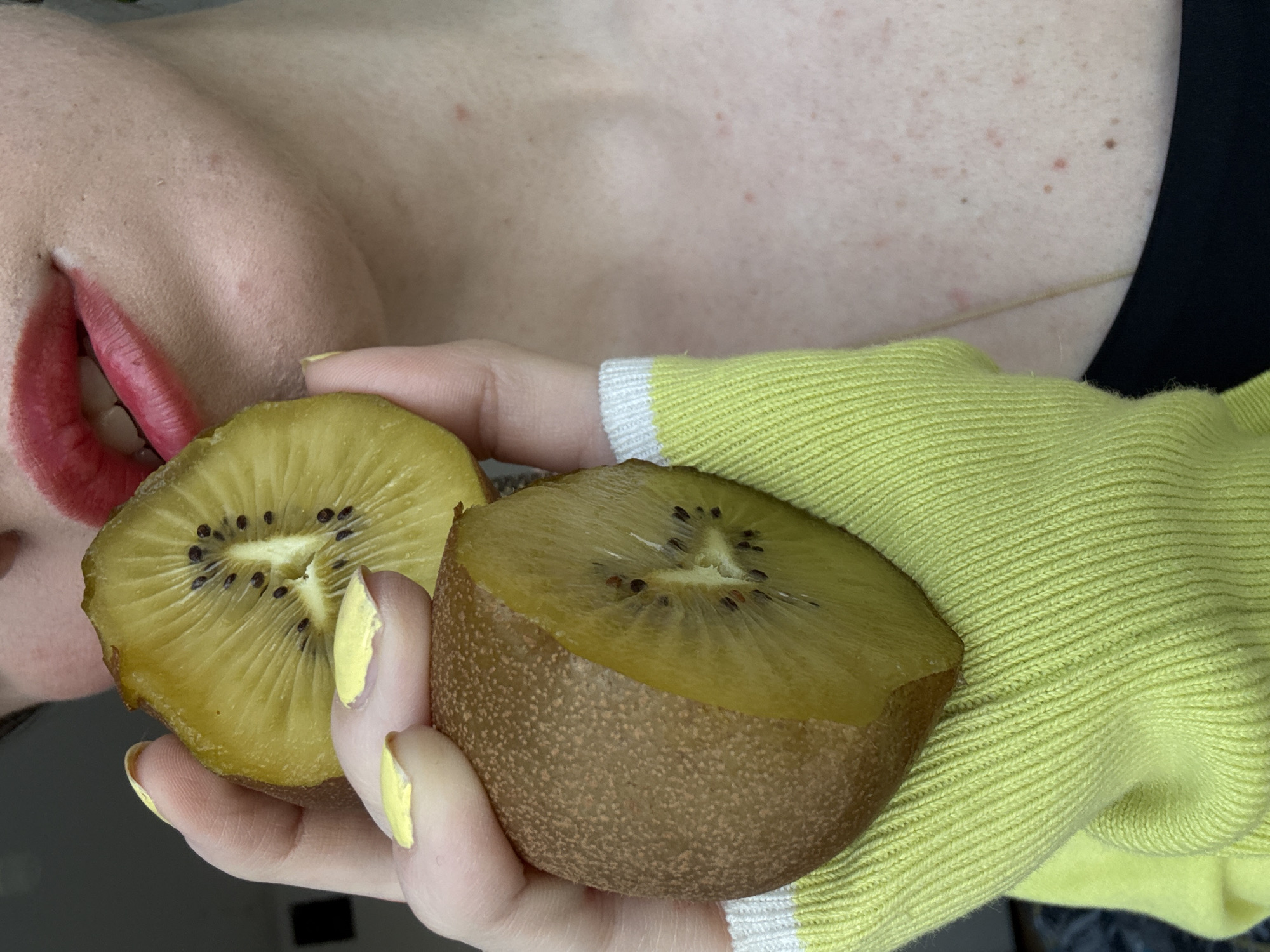A close-up, eye-level shot shows a person holding two halves of a golden kiwi fruit in their hands. The person has red lipstick and yellow nail polish. The person is wearing a light green sweater with white stripes on the cuff. Part of the person's chest and neck are visible. The background is out of focus. The location is not discernible, and there is no indication of Moscow, Russia.
