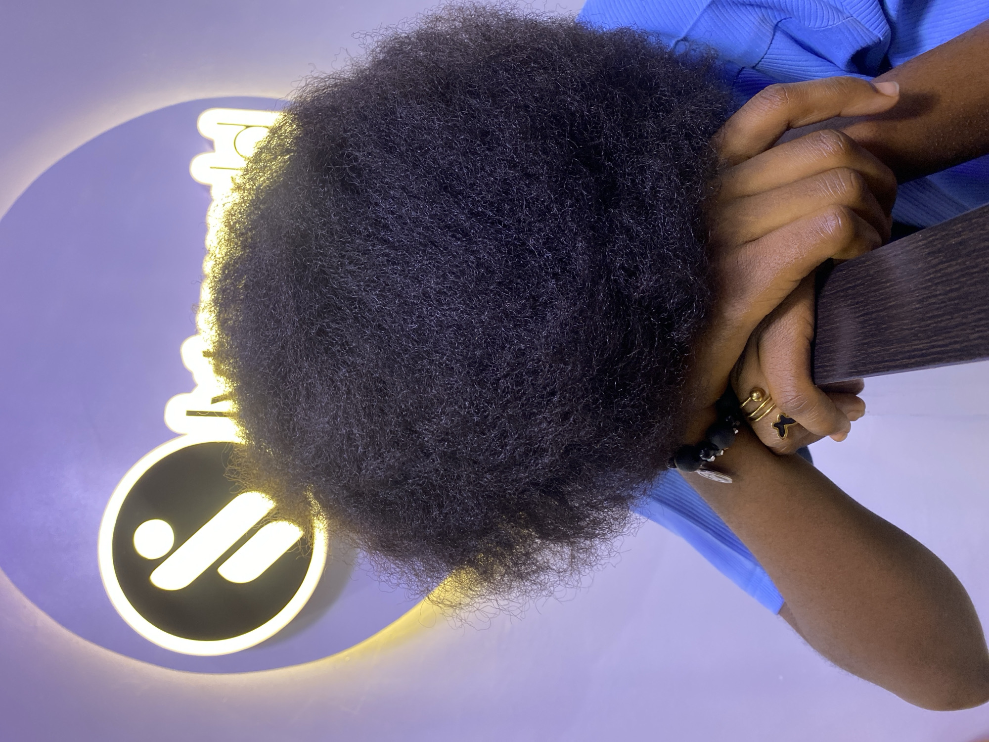 The image shows a close-up of a person with a large afro hairstyle, leaning their head on their arms. The person is wearing a blue collared shirt, a black beaded bracelet, and rings on their fingers. Behind them, a circular logo with a stylized "i" symbol illuminated by yellow light is visible against a purple background. The logo is partially obscured by the person's hair. The lighting suggests an indoor environment, possibly a studio or an office space, with a soft purple ambient glow. The overall impression is one of contemplation or rest, with the person's face hidden from view. The image does not provide any specific information that would allow for the identification of the location as Gura Top, Nigeria.