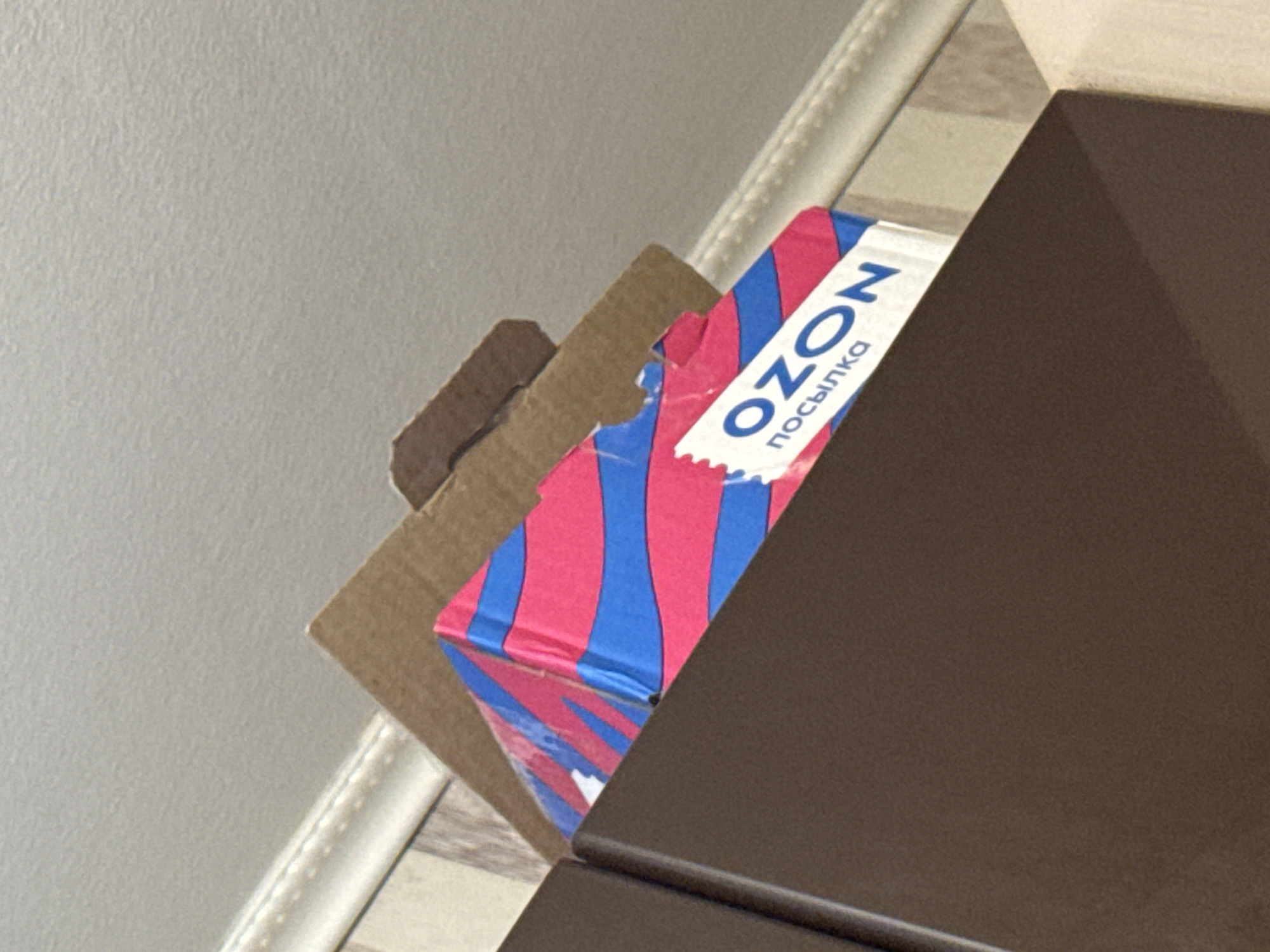 A partially visible cardboard box with the Russian e-commerce company OZON's logo and the word "посылка" (parcel) in blue text on a white label with a pink and blue striped design is partially obscured by a brown surface. The box appears to be resting on or near a light-colored surface, possibly a table or shelf, next to a white wall and decorative moulding. The photograph's perspective is from a slightly elevated angle. The location is likely Novo-Nikol'skoe, Russia, based on the prevalence of OZON deliveries there.