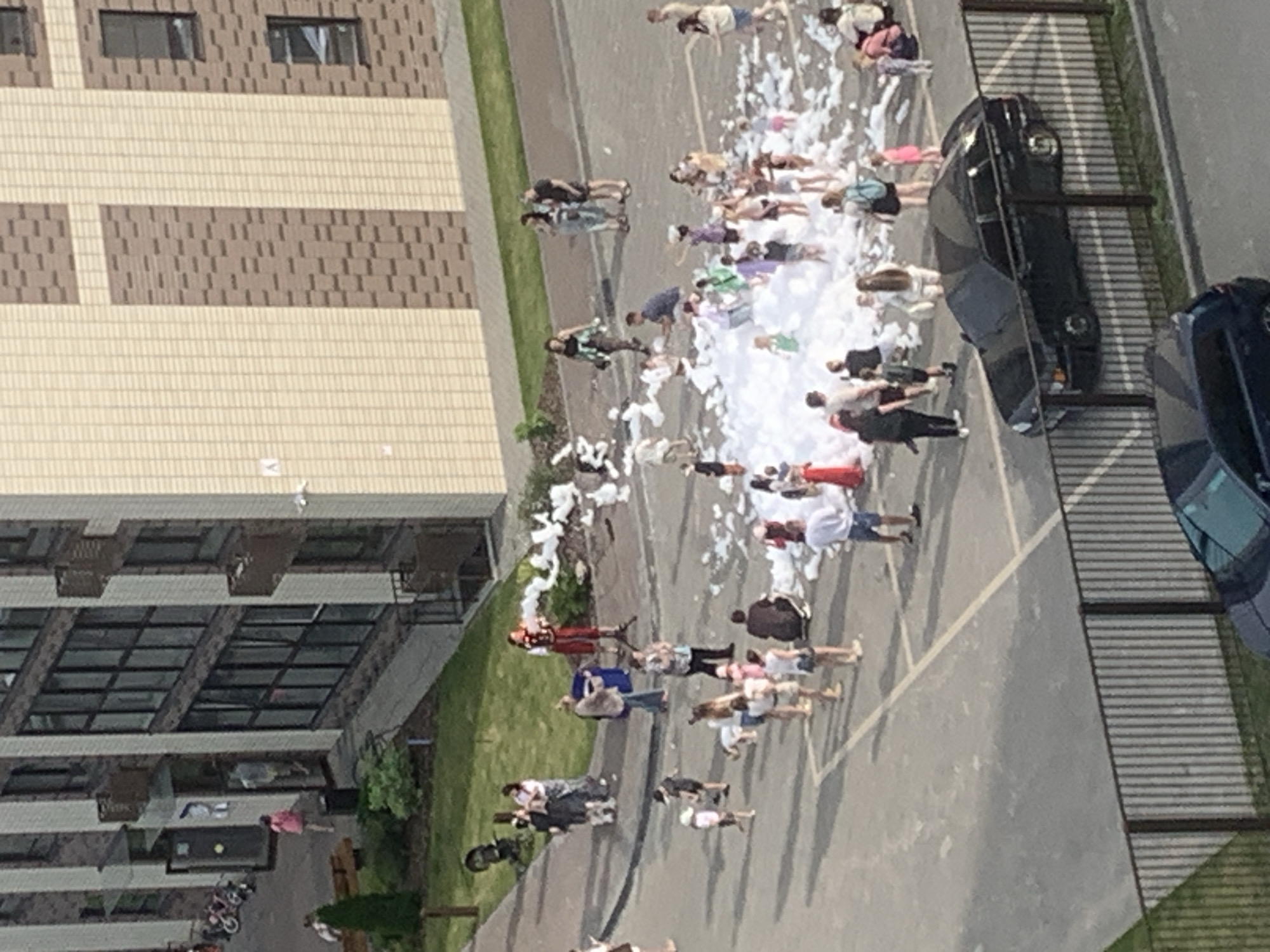 The image depicts a residential courtyard in Kirov, Russia, likely on a summer day, as evidenced by the light clothing worn by people and the sunny conditions. The scene is dominated by a large pile of foam, suggesting a foam party or event. Approximately 30-40 individuals, including children and adults, are gathered around the foam, with some actively playing in it and others observing from the periphery.
A modern, multi-story apartment building with light-colored and brick facades forms the backdrop. The ground surface is asphalt, marked with parking spaces. Several cars are visible, including a black sedan parked in a marked bay and a partial view of another vehicle. Fencing separates the courtyard from a grassy area.
In the background, near the building entrance, a few children are seen with bicycles and a stroller, indicating a family-friendly environment. Two adults in festive attire are positioned near the foam area, possibly responsible for managing the foam machine or entertaining guests. The overall atmosphere appears to be one of recreation and community gathering.