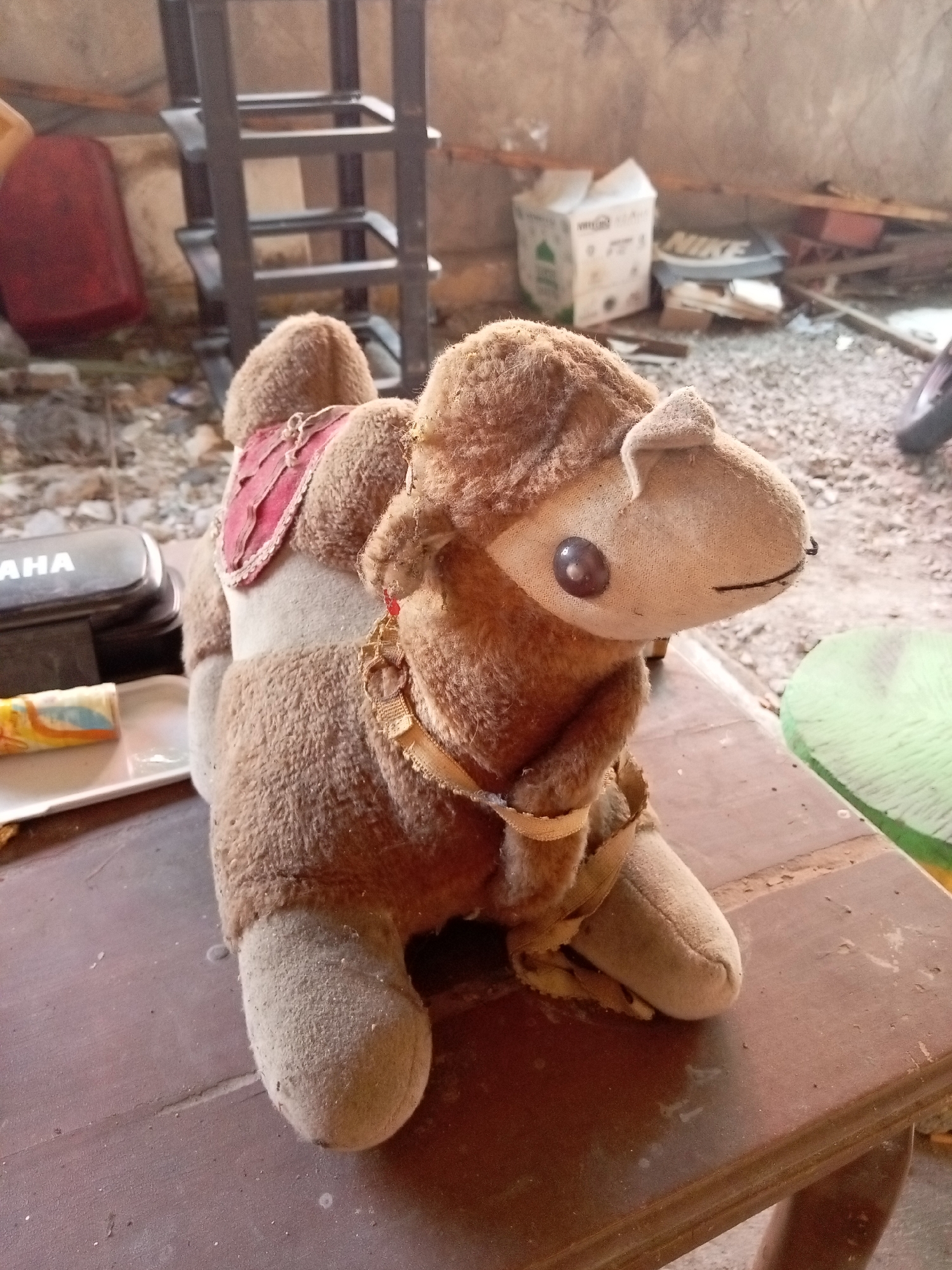 The image depicts a plush camel toy sitting on a dark brown wooden table. The toy is light brown with a red saddle. In the background, there are various objects including a red container, a metal shelf unit, a white box with green accents, and a discarded Nike shoe box. The ground appears to be gravel. A partially visible green circular object is present to the right of the table. The overall setting suggests an outdoor or semi-outdoor area, possibly a storage space or a yard in Mojokerto, Indonesia.