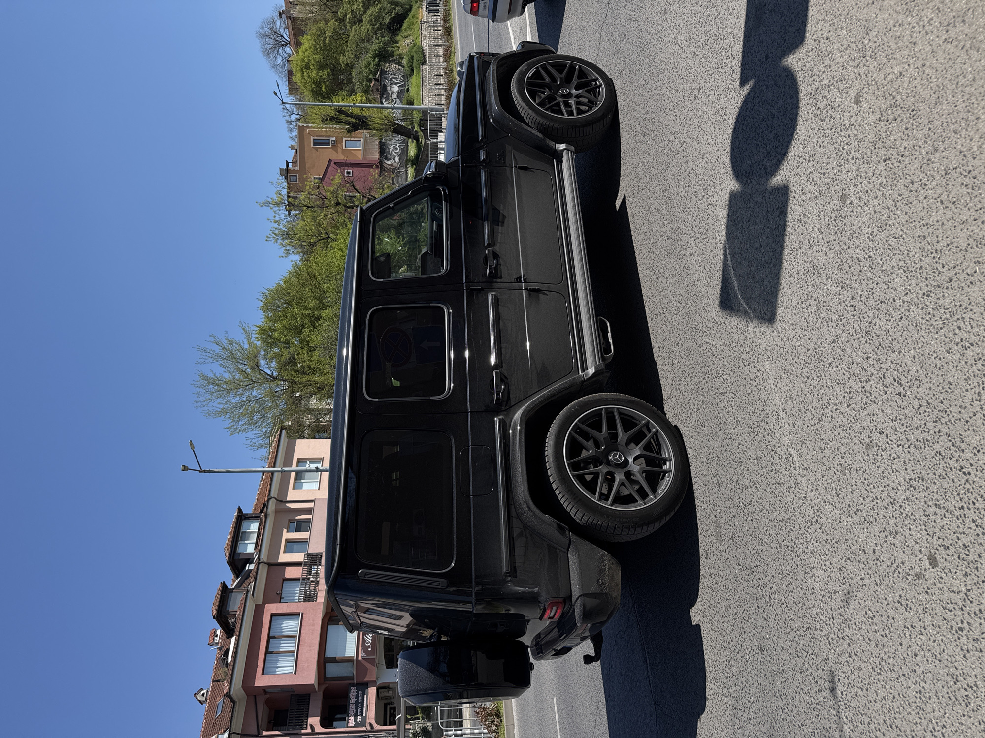 A black Mercedes-Benz G-Class AMG SUV is parked on the side of a paved road in Plovdiv, Bulgaria. The SUV is oriented with the rear towards the lower left corner of the frame. The rear features a spare tire mounted on the tailgate. The vehicle is parked on the right side of the road, with the background revealing residential buildings with pink and beige facades. A row of trees lines the road further back. The sky is clear and blue. Shadows are cast on the road surface by an unidentifiable object. The wheels have AMG-specific rims. There are no visible people in the frame.