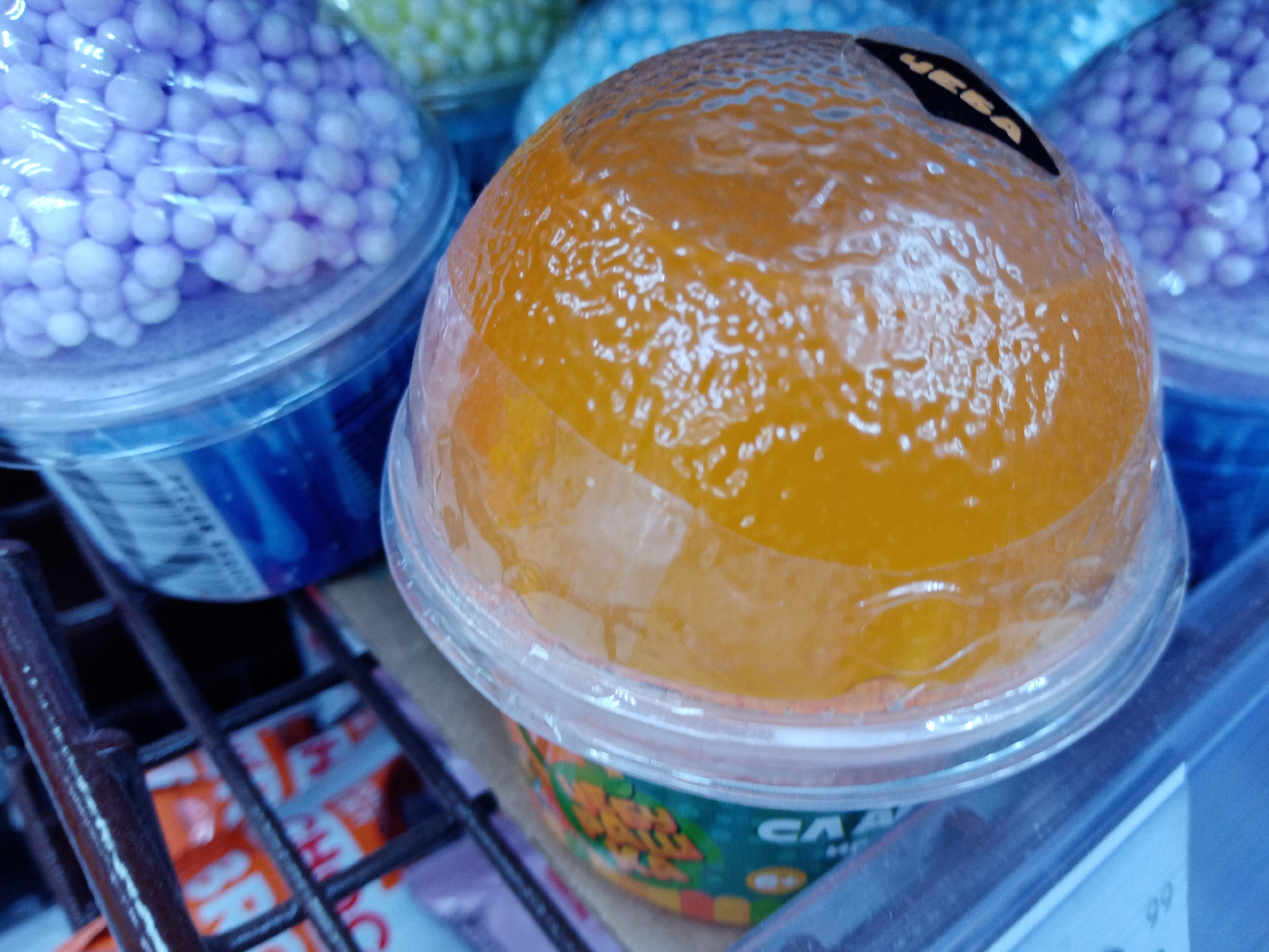 The image captures a close-up view of several transparent plastic containers on a shelf, likely in a retail store in Elektrostal, Russia. The most prominent container in the foreground is filled with a vibrant orange, viscous, gel-like substance, possibly a form of slime or jelly, which has a textured and glossy surface. A small, black, irregularly shaped sticker with the word "IKEA" (upside down) is adhered to the top of this container. Its side label, partially visible, displays Cyrillic text including "СЛАЙМ" (SLIME) and "СА".
To the left and slightly behind the central orange container, another identical container holds numerous small, light purple, spherical beads, likely foam beads used in crafts like slime. Further in the background, containers with yellow and blue beads are also visible. The containers rest on or next to a dark metal grid-like shelf or display rack. A white tag with the numbers "99" is visible on the far right, possibly indicating a price. The scene suggests an indoor environment with artificial lighting, typical of a store selling toys, crafts, or novelties. No human activity is depicted; the image focuses on the products on display.