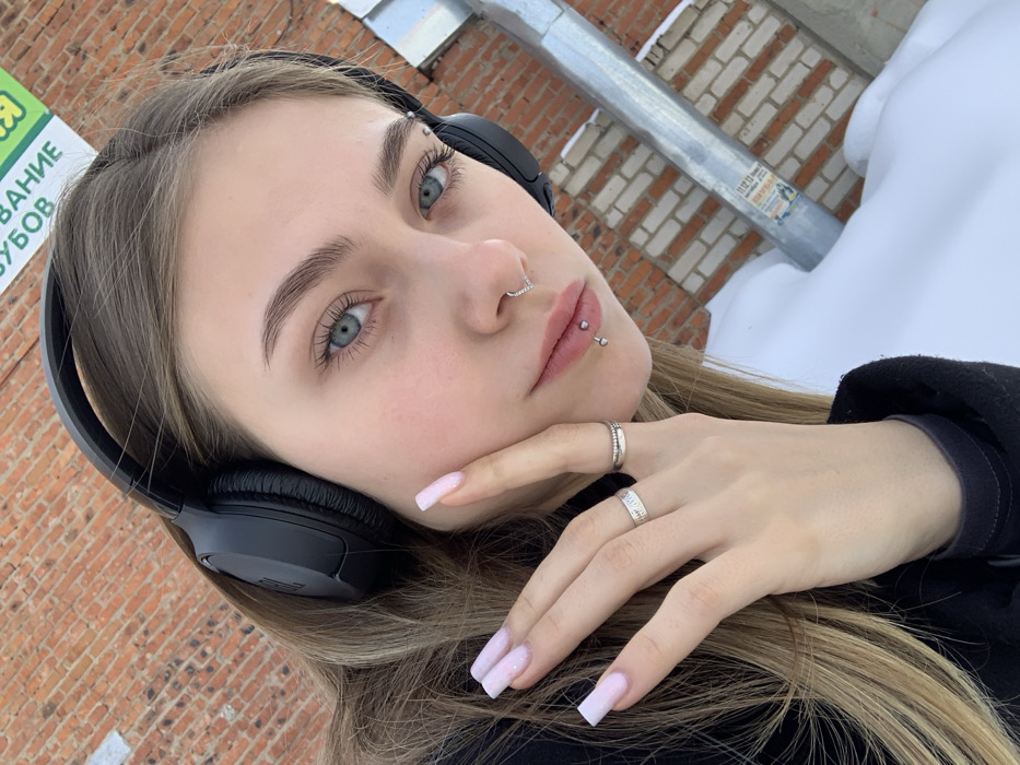 A young female is depicted outdoors in Chaikovsky, Russia, within an urban environment characterized by a brick building and snow cover. The subject, a female with light blue eyes and light brown hair, wears black JBL over-ear headphones. Her right hand, featuring long, coffin-shaped nails painted with light pink glitter polish, is positioned with fingers splayed, lightly touching her right cheek. She wears two silver-toned rings on her right ring finger: a thin band and a wider band with an etched pattern. Facial piercings include a V-shaped septal ring with small gems and a vertical labret piercing on her lower lip with a barbell. Her gaze is directed towards the camera. She is dressed in dark outerwear, likely a coat or jacket. The background consists of a reddish-brown brick wall. A partial white and green sign, displaying Cyrillic text including "ЗУБОВ" (teeth/dental), is attached to the upper section of the brick wall. A corrugated metal downspout runs vertically along the right side of the wall, featuring remnants of attached papers. The ground in the foreground and along the building's base is covered by a significant layer of undisturbed white snow.