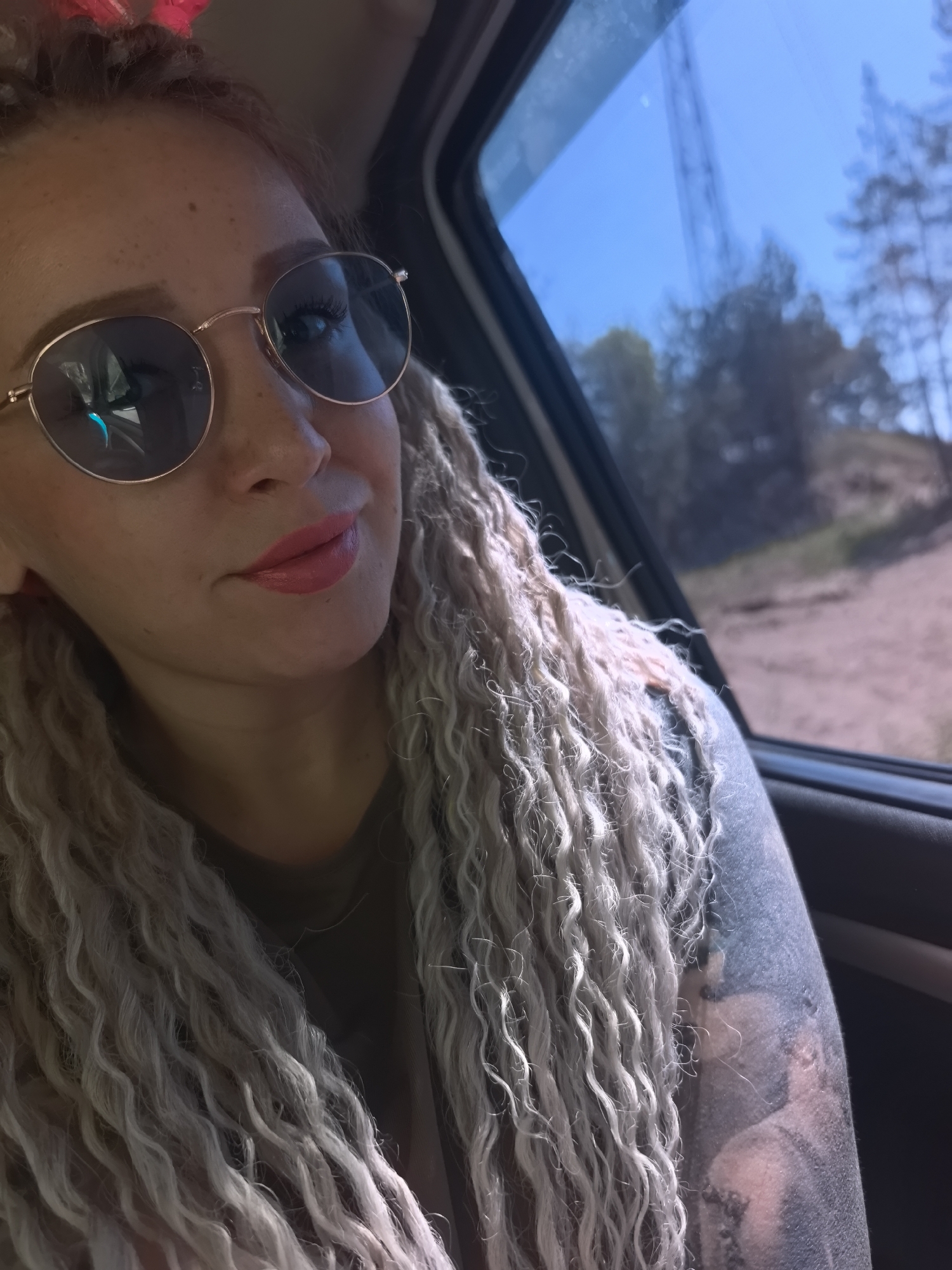 A woman with blonde, curly hair and round sunglasses is sitting in the passenger seat of a vehicle. She has tattoos on her left arm, and is wearing lipstick. Outside the car window, trees and a power line are visible. The location is potentially near Noviy, Russia, based on visual cues of the environment and potential assumptions from the prompt context, but this is not confirmed within the image.