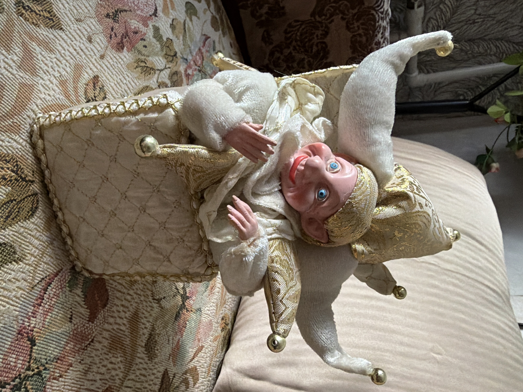 A close-up, eye-level shot shows a vintage jester figurine sitting inside a quilted box. The figurine has a pink, smiling face with bright blue eyes and pointed ears. It wears a white and gold jester costume with a floppy hat adorned with gold trim and small gold bells. The figurine's hands are raised, palms facing outwards. The box the figurine is in is made of quilted fabric with gold trim and a diamond pattern. The figurine and box are placed on a patterned fabric surface, likely a couch or chair, with floral motifs in muted colors. In the background, a blurred section of a wall with a textured pattern and a plant are visible. The scene does not appear to be located in Nizhny Novgorod, Russia, as there are no discernible landmarks or contextual elements to suggest this location. The lighting is natural and appears to be coming from the side.