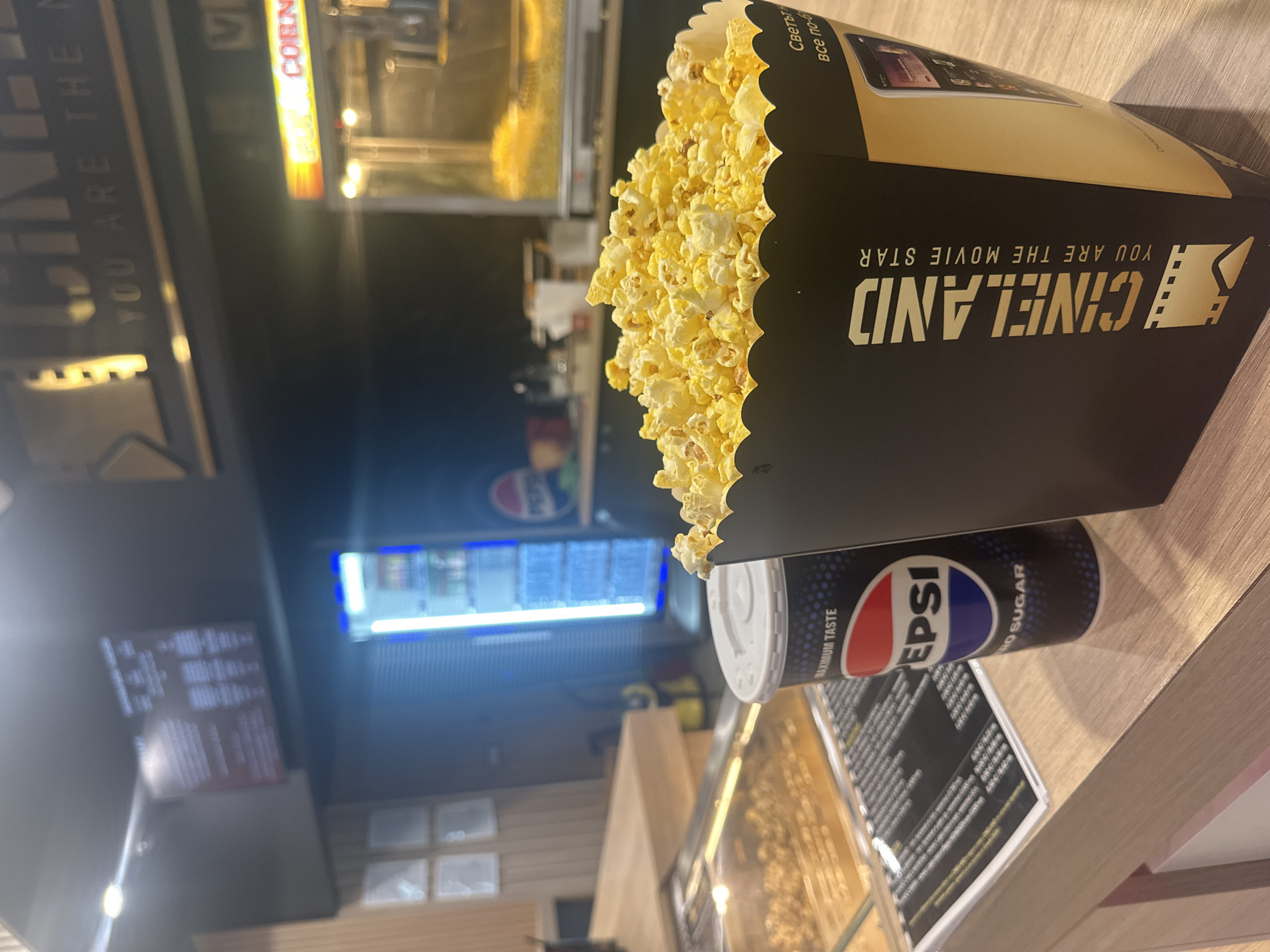 The image shows a large container of Cineland popcorn and a can of Pepsi Zero Sugar on a light brown wooden surface. In the background, there is a partially visible popcorn machine and a menu board. "CINELAND YOU ARE THE MOVIE STAR" is printed on the side of the popcorn container. "MAXIMUM TASTE EPSI ZERO SUGAR" is visible on the Pepsi can. The scene is likely within a Cineland cinema. Given the Cyrillic text on the popcorn box ("Светьт все по-б"), the image is likely taken in a Cineland cinema in Sofia, Bulgaria.