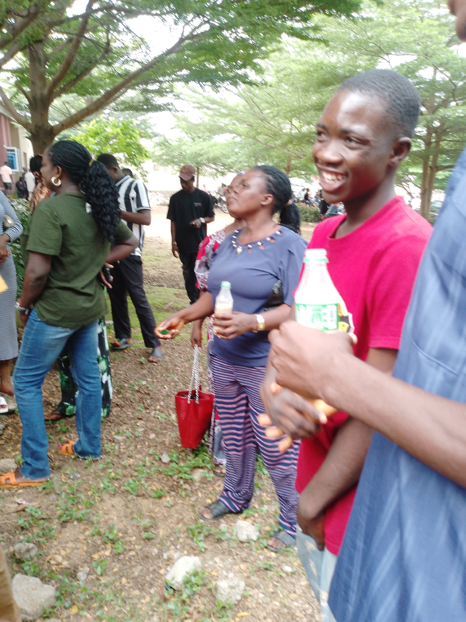 In Akwanga, Nigeria, a group of people are gathered outdoors, possibly at an event. The foreground is dominated by a young man wearing a bright red t-shirt, smiling broadly as he looks to his left. He is holding a bottle of drink. To his left, a woman in a dark blue top and striped pants also holds a bottle of drink. Behind them, a woman in a green shirt and blue jeans stands with her hand on her hip, facing away from the camera. Another man in a black t-shirt and dark pants is visible further back, and a person in a blue shirt is seen in the far right of the frame. The ground is covered with dirt and sparse vegetation, and trees with lush green leaves form a canopy overhead, suggesting a daytime setting in a park or outdoor venue.