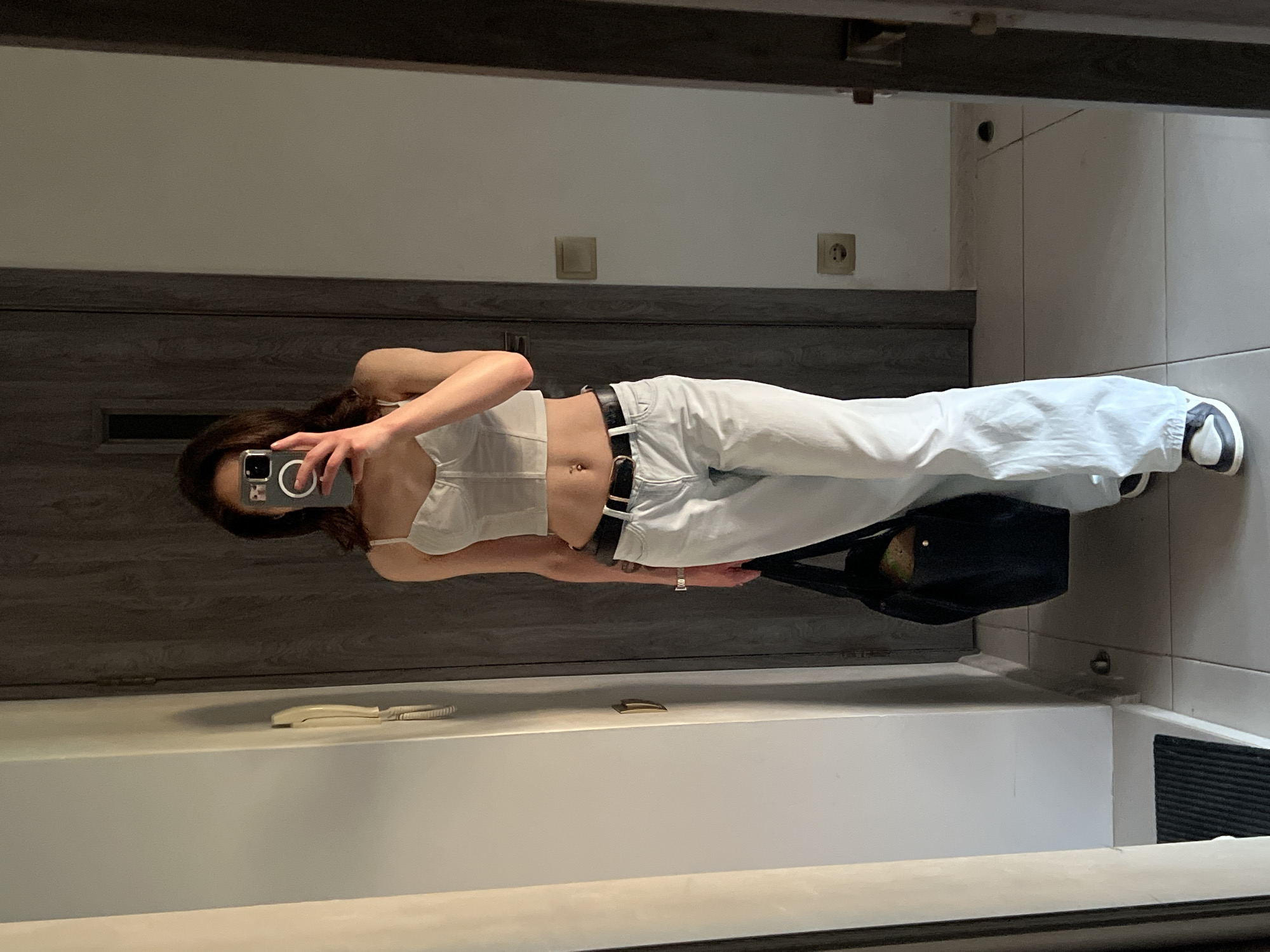 A young woman, with dark hair, stands in an indoor hallway and takes a mirror selfie. She is wearing a white corset top, light-colored baggy jeans, a black belt, and black and white sneakers. She is holding a black tote bag in her left hand. The hallway features a light-colored tiled floor, light-colored walls, and a dark wooden door. A white intercom is mounted on the wall to the left. The scene appears to be in an apartment building hallway.
