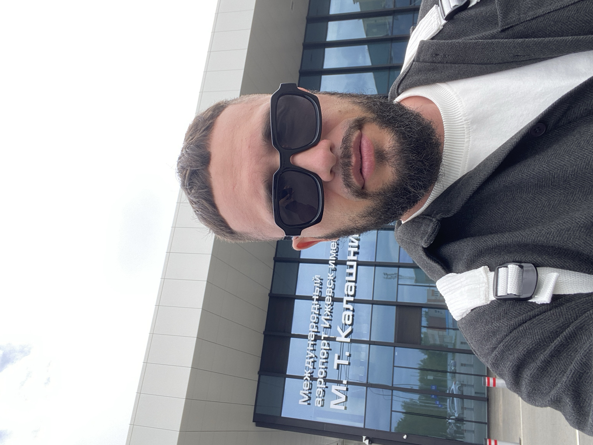 A man with a beard and sunglasses stands in front of the Izhevsk Airport named after M. T. Kalashnikov. The airport is a modern building with large glass windows. The man is wearing a dark jacket over a white sweater. He has a white strap across his chest, suggesting he is carrying a bag. The sky is cloudy.