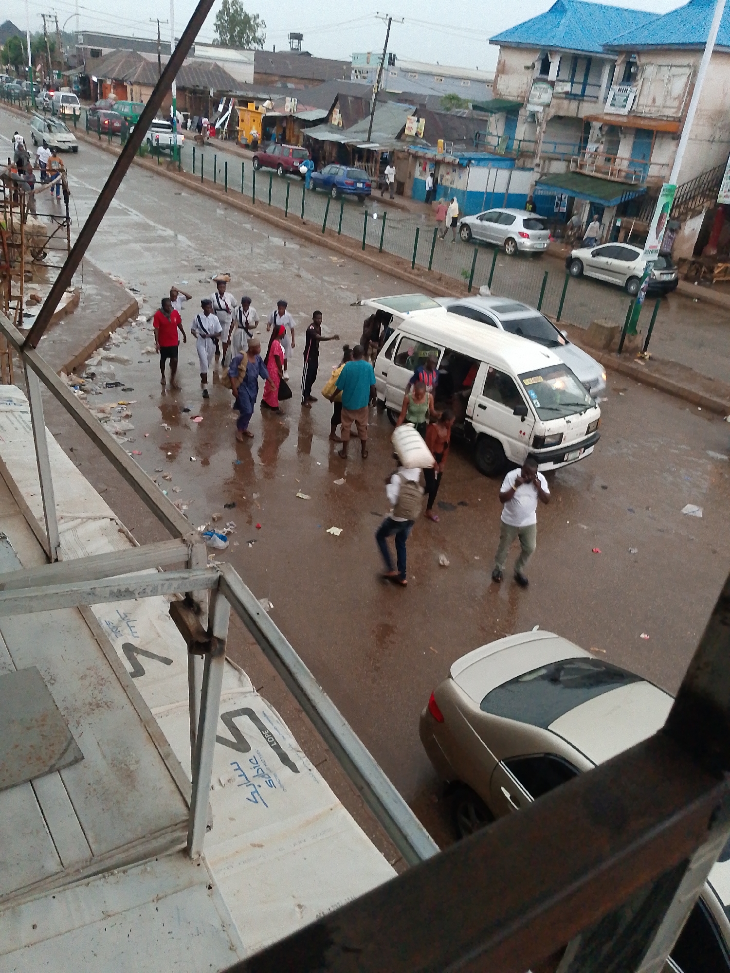The image depicts a street scene in Bukuru, Nigeria. The street is wet and littered with debris. Several people are walking on the street, including a group of individuals wearing white clothing with what appear to be white caps and sashes, a person wearing a red shirt and black shorts, and others in casual attire. A white van is parked on the street and several individuals are either boarding or exiting the vehicle. Two sedans, one beige and one silver, are also parked on the street. Buildings line the street, with a mix of architectural styles and materials, including some with blue roofs and others with corrugated metal roofs. There are also various commercial establishments visible along the street. The scene is viewed from an elevated position, with structural elements of the building visible in the foreground.