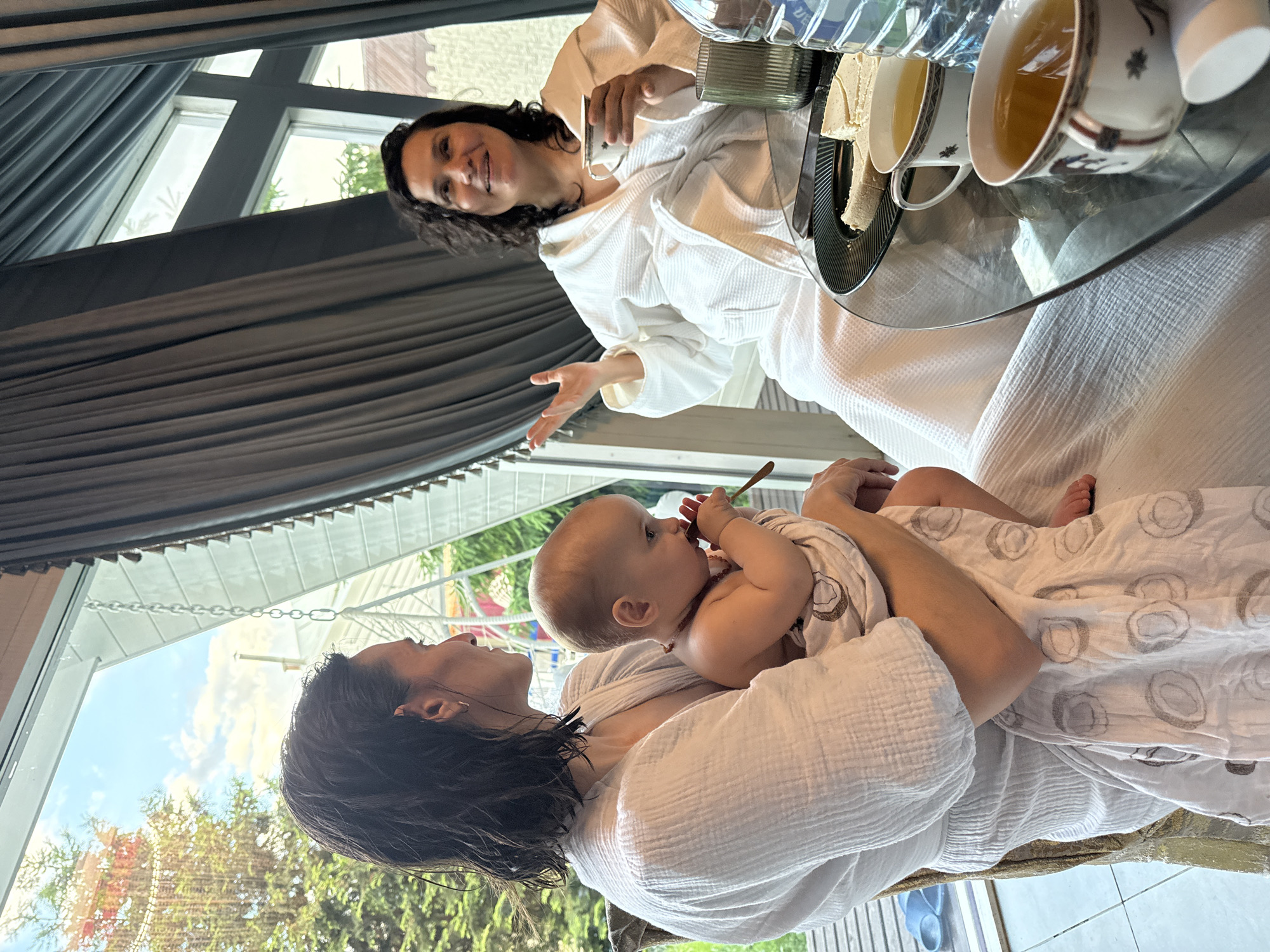A woman in a white bathrobe holds a baby in her arms, while a second woman in a white bathrobe sits at a table. The baby holds a small wooden spoon in its mouth. The table holds two cups of tea, a container, a tray with a white square of food, and two water bottles. A window behind the seated woman shows a gray curtain and a grid of window panes. A window behind the standing woman reveals green trees, a blue sky, and a white overhang. The location is indoors. The city of Чёрное, Russia, is not directly discernible from the image.