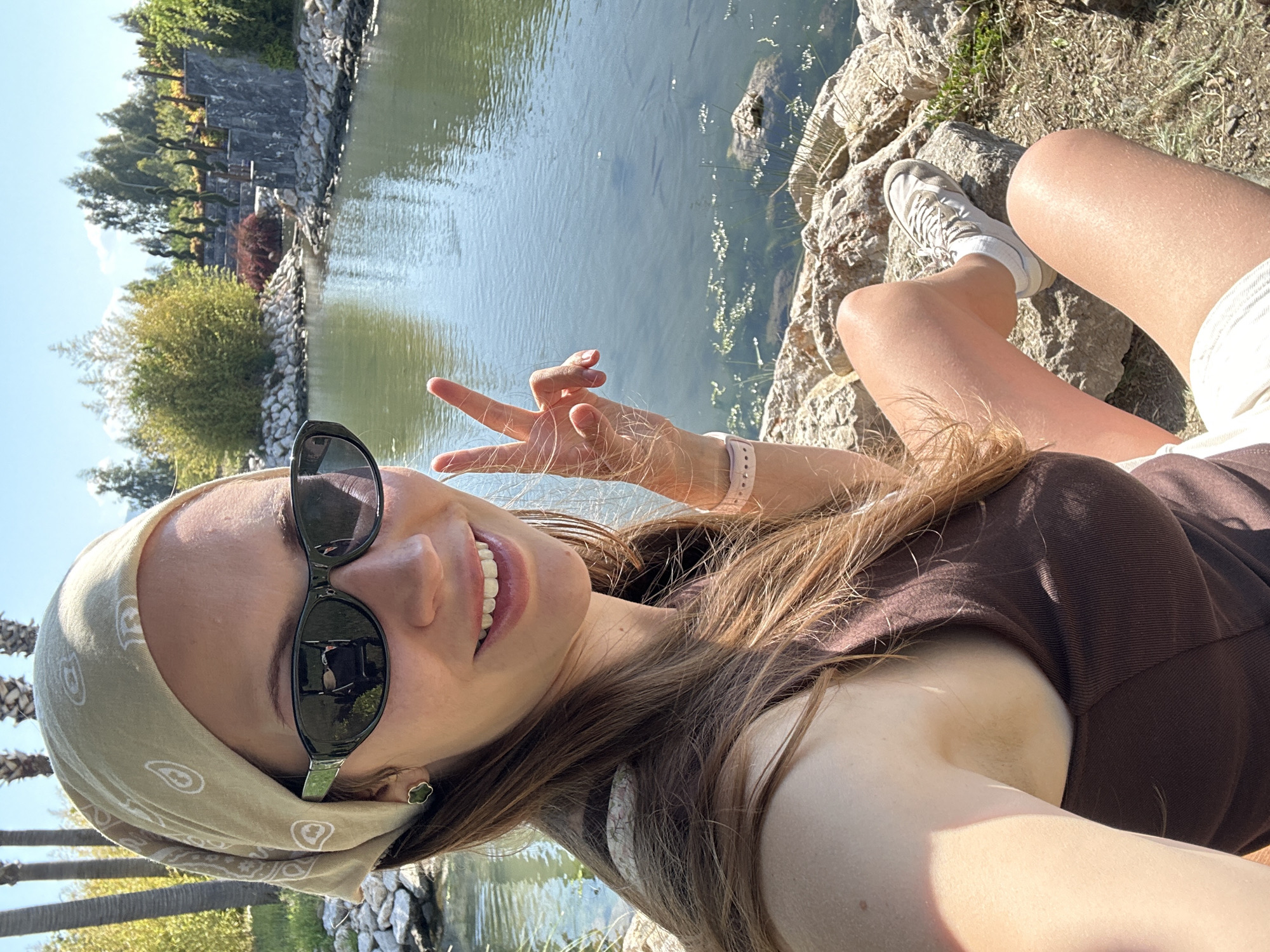 A young woman is captured in a sunlit selfie, taken outdoors beside a body of water. The image is rotated 90 degrees clockwise. She is positioned on a rocky bank, facing the camera with a wide smile and making a peace sign with her left hand. Her long, light brown hair flows down her shoulders. She wears dark sunglasses that reflect the surrounding environment, a light beige bandana with a paisley pattern around her head, a brown top, light-colored shorts, and white sneakers. A light pink smartwatch or band is visible on her wrist.
The background features a calm body of water, likely a lake or pond, reflecting the clear blue sky. Across the water, there's a lush green landscape with various trees and shrubs, including palm trees on the left. A distinctive tiered stone structure or wall is visible on the far bank, partially obscured by greenery, suggesting a landscaped area. The bright sunshine indicates it is daytime, likely mid-day, with clear weather. The woman's smile and peace sign convey a cheerful and relaxed mood. No visible text is present in the image. The setting suggests a park or recreational area in Fethiye, Turkey.