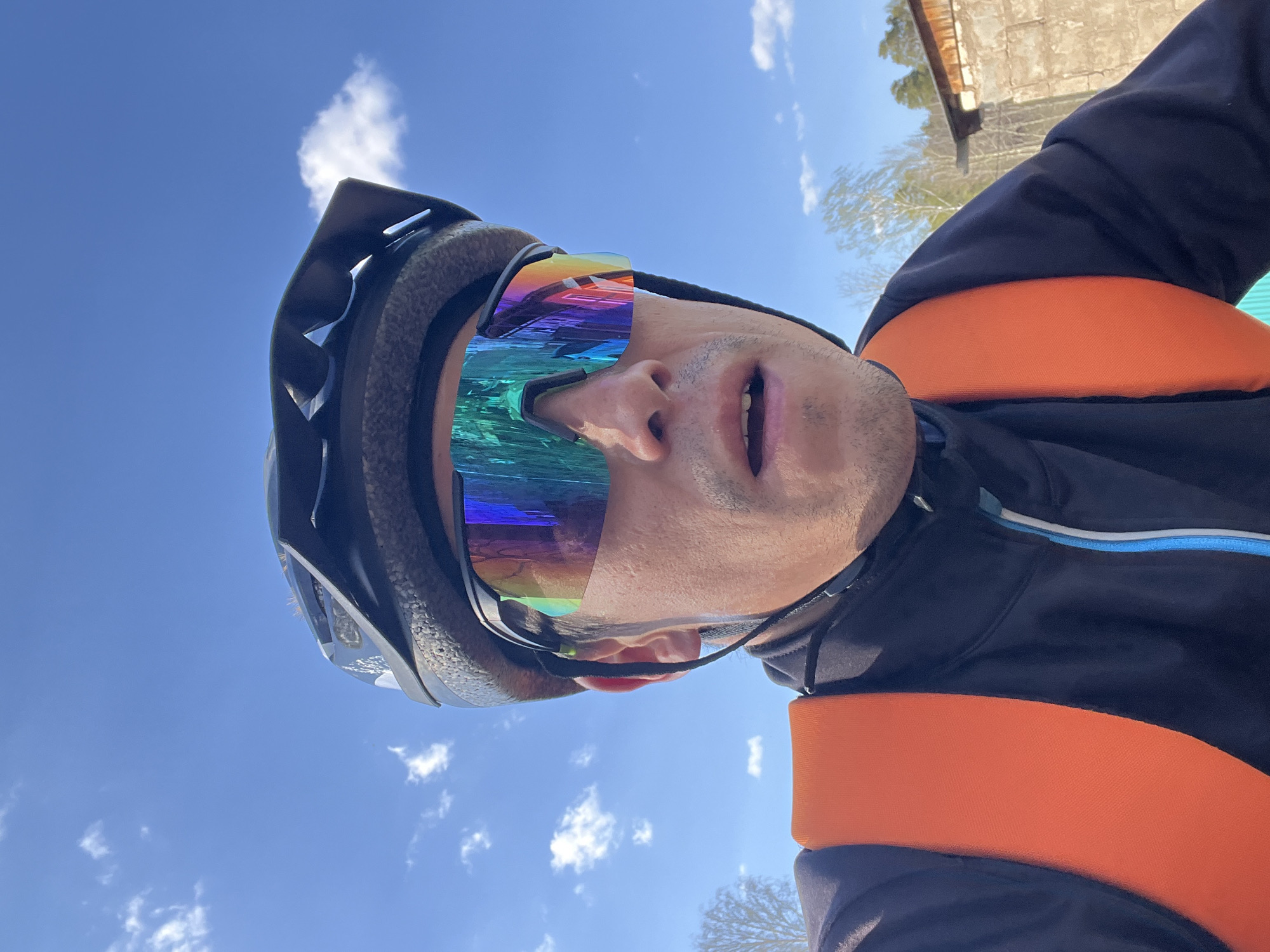A low-angle, outdoor selfie shows a person wearing a dark-colored helmet and iridescent, rectangular sunglasses, with the sky visible behind them. The person is also wearing a black jacket with orange straps or a vest. Some trees and a building are slightly visible in the background. The picture was taken in the city of Chaikovsky, Russia. The person appears to be looking up at the camera.
