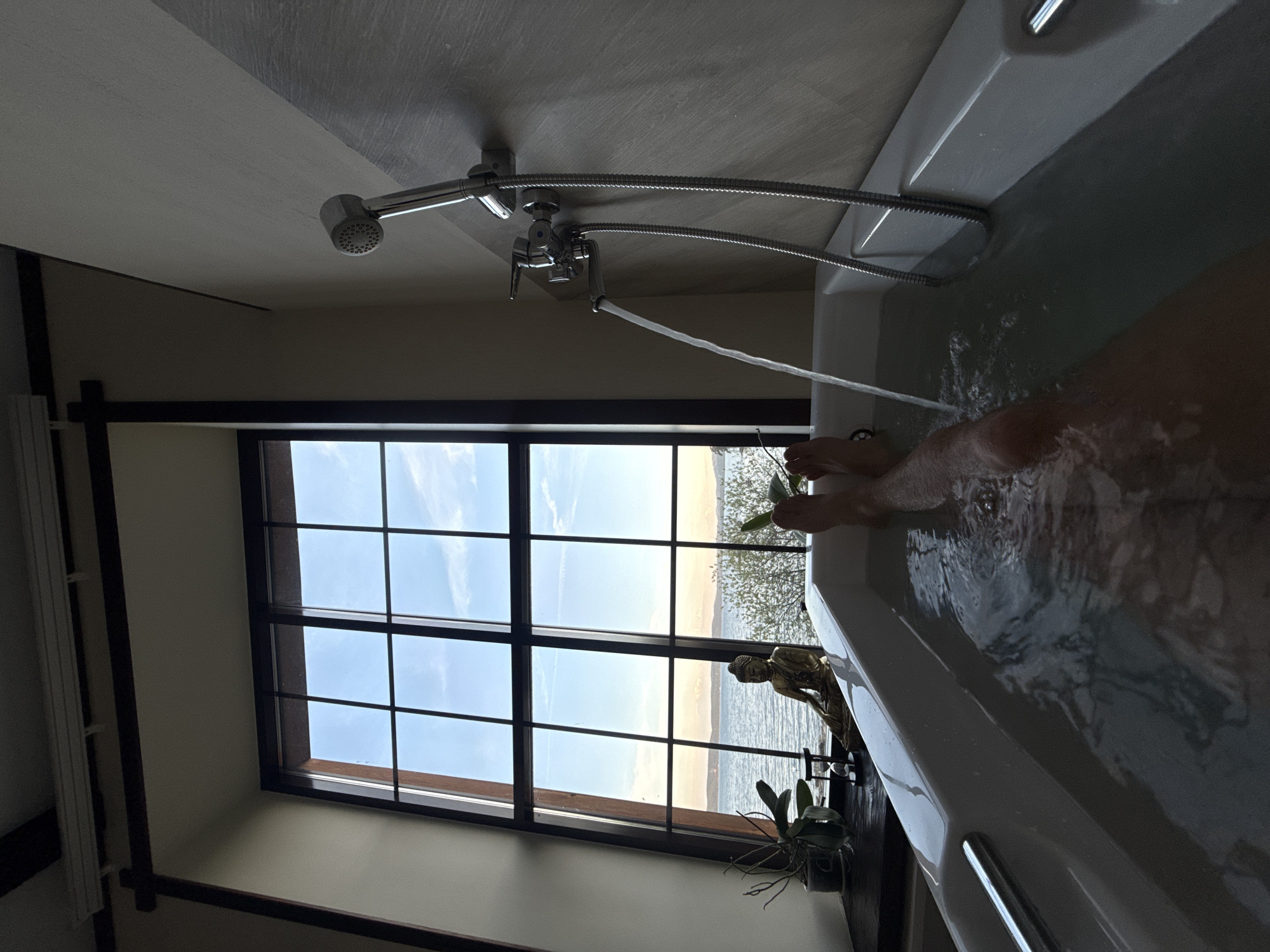 The image shows a person in a bathtub, likely located in or near Zolotoi Plyazh, Russia, given the view. The tub is partially filled with water, and the person's legs and feet are visible in the water. A showerhead is mounted above the tub, and water is running from the faucet into the tub. A large window is visible, offering a view of the sky, a beach, and the sea. A statue of Buddha and a potted plant are visible on the windowsill, next to the tub. The room appears to have a modern architectural style, with dark wooden accents on the window frame.