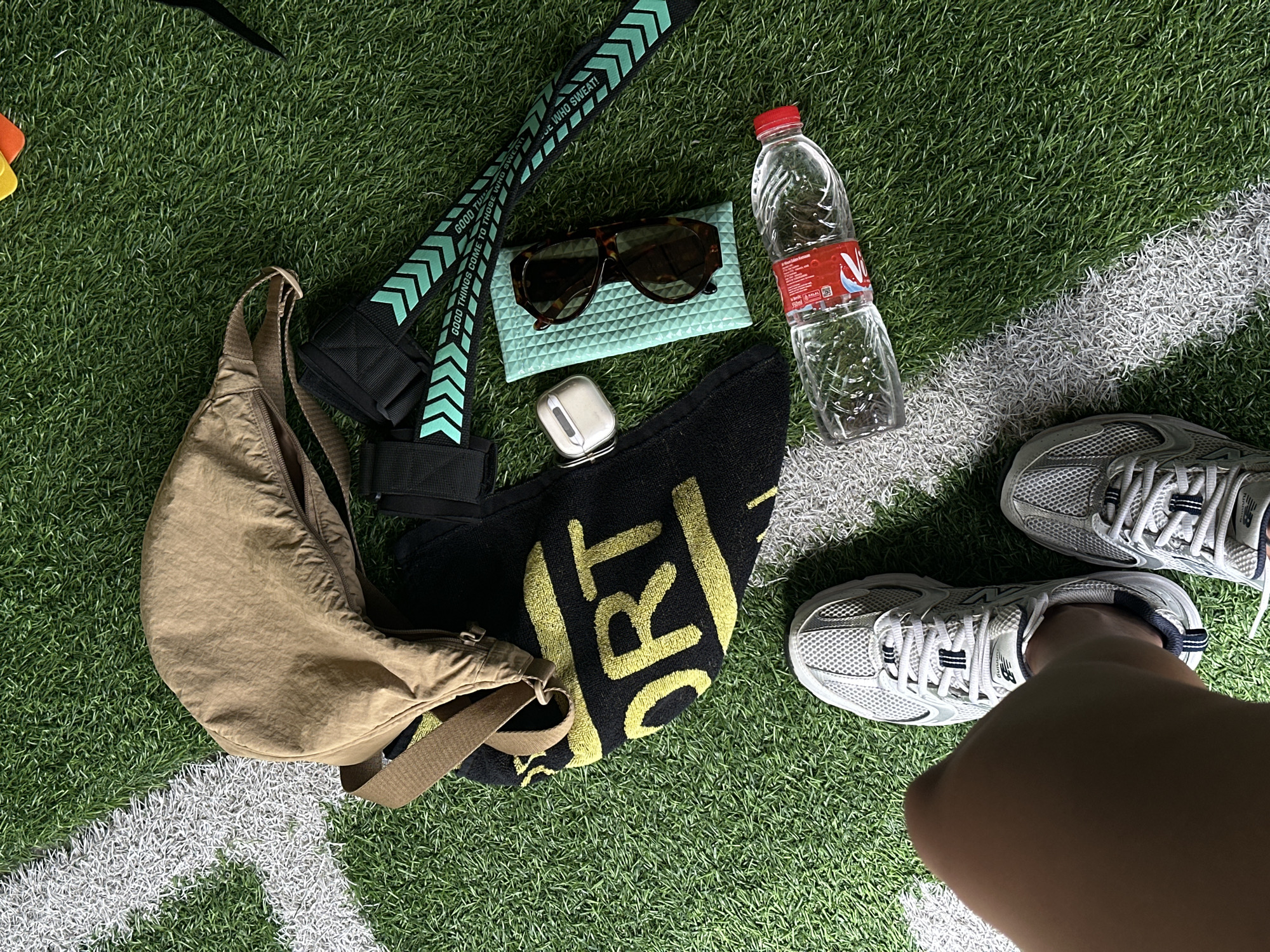 The image shows a flat lay of sports and personal items arranged on an artificial turf surface, marked with white lines, suggesting a sports field or court. The items include a beige crossbody bag, a black towel with yellow lettering that appears to spell "SPORT," a pair of grey and white athletic shoes, a water bottle with a red cap and label, a pair of tortoiseshell sunglasses in a light blue textured case, and white wireless earbuds. Also visible are two black resistance bands with a teal chevron pattern and text that reads "GOOD THINGS COME TO THOSE WHO SWEAT!". The lower left corner shows a portion of a person's legs and feet wearing the athletic shoes. The setting appears to be outdoors, possibly in North Kuta, Indonesia, given the artificial turf and the presence of sports equipment.