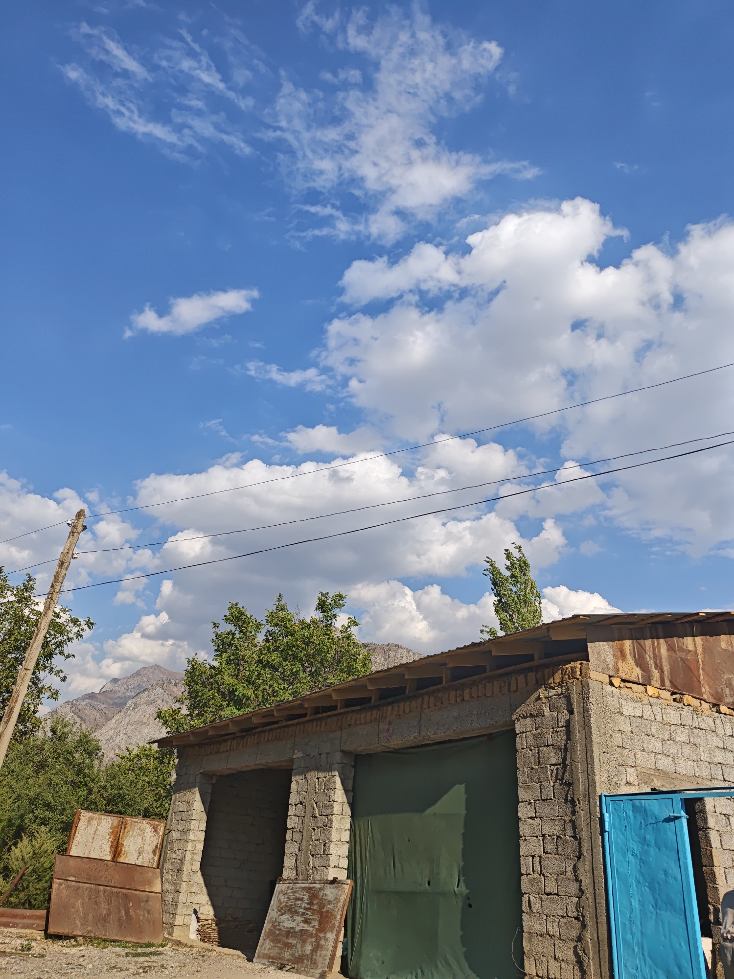 The image depicts an outdoor scene with a clear blue sky dotted with white clouds. In the foreground, a building constructed of grey bricks with a corrugated metal roof is visible. The building appears to be unfinished or in disrepair, with exposed wooden beams supporting the roof. There are large green tarps covering openings in the building, suggesting they are being used as makeshift doors or covers. To the left of the building, two large, rusty metal sheets are propped up. A wooden utility pole with insulators stands to the left of the building, with power lines stretching across the frame. Behind the building, lush green trees partially obscure a mountainous landscape. The overall impression is of a rural or semi-urban environment, possibly in a developing area, given the building materials and the utilitarian nature of the structures. The setting is likely the city of Yakkatut, Uzbekistan. There are no people or animals visible in the image.