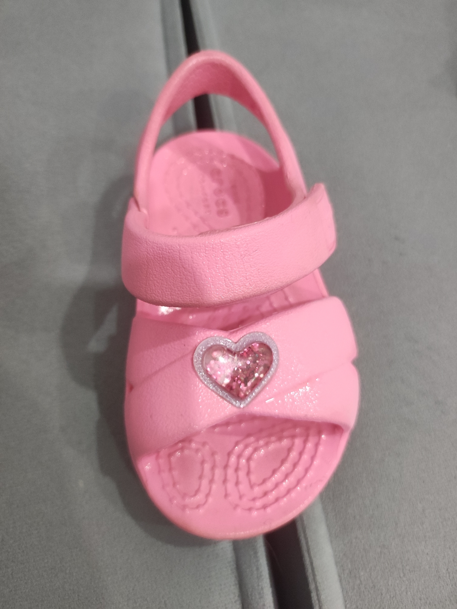 A close-up shot displays a single pink Crocs sandal, designed for children, featuring a heart-shaped embellishment filled with glitter. The sandal rests on a gray surface, likely a piece of furniture, possibly a couch. The image lacks any details suggesting a specific location in Votkinsk, Russia, or any indication of people, actions, or interactions. The environment is limited to the immediate surface on which the sandal is placed.