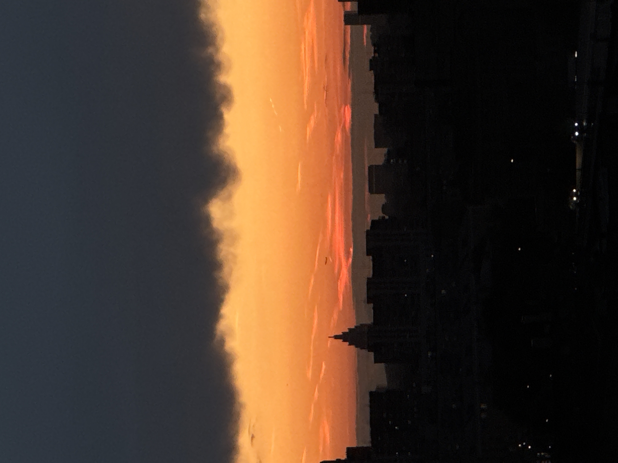 This image captures a dramatic sunset over a cityscape, likely Moscow, Russia, given the architectural style and general urban density. The sky is dominated by a gradient of intense orange and yellow hues near the horizon, transitioning to a dark, brooding grey cloud mass at the top. A distinct silhouette of buildings forms the lower half of the frame, with a prominent, stepped tower structure rising above the other structures, resembling Stalinist architecture often found in Moscow. Tiny lights dot the darkness of the foreground, indicating city lights and possibly traffic on a road or highway. The overall impression is one of an urban environment at dusk, with the natural spectacle of the sunset providing a striking contrast to the man-made structures.