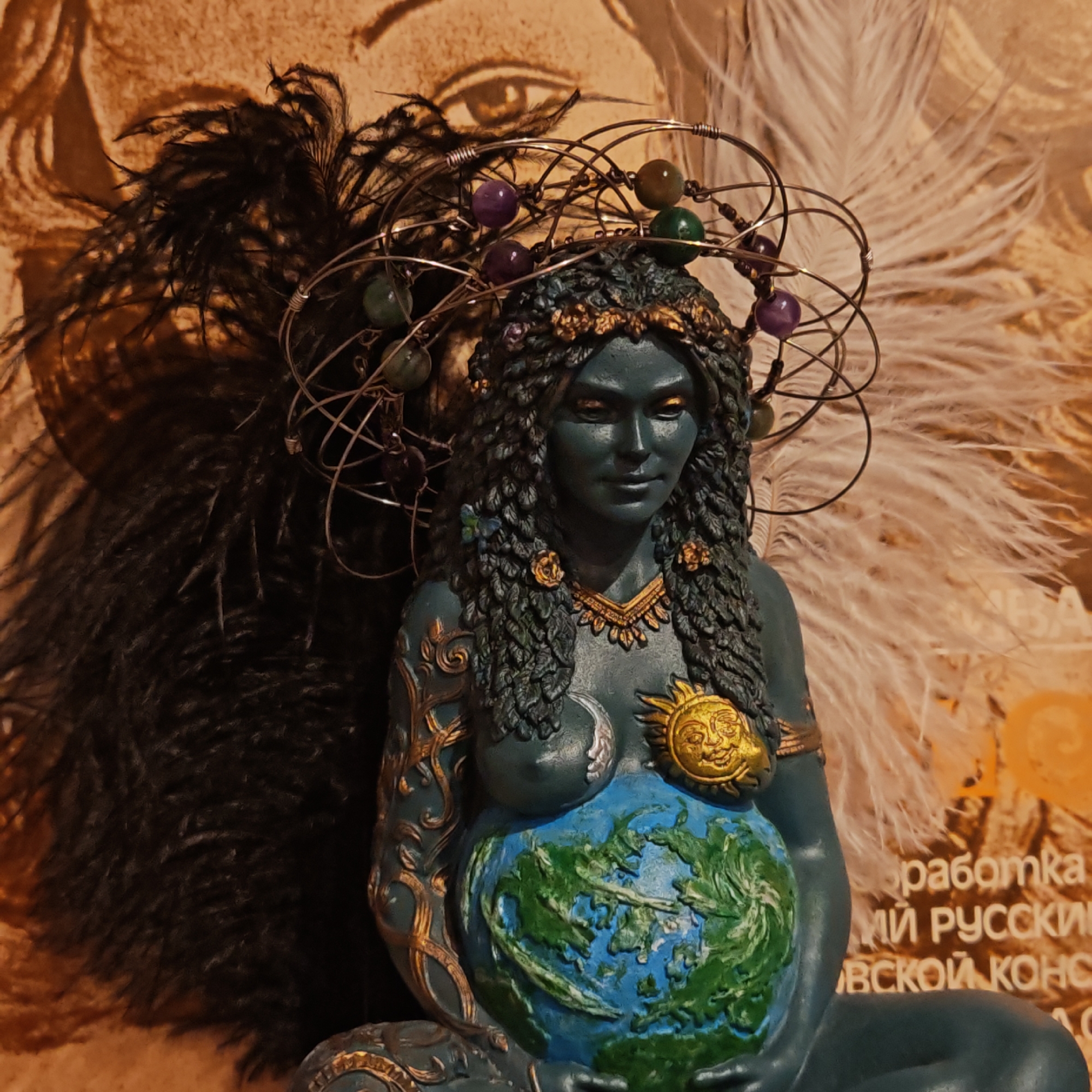 A statue of a blue-skinned female figure with a wire halo and beads sits in front of a backdrop of feathers and blurred text. The figure's belly is painted like the Earth. Her left breast is adorned with a moon and her right with a sun. She wears a crown and a necklace, and her left arm is decorated with gold filigree. The location is Novo-Nikol'skoe, Russia.