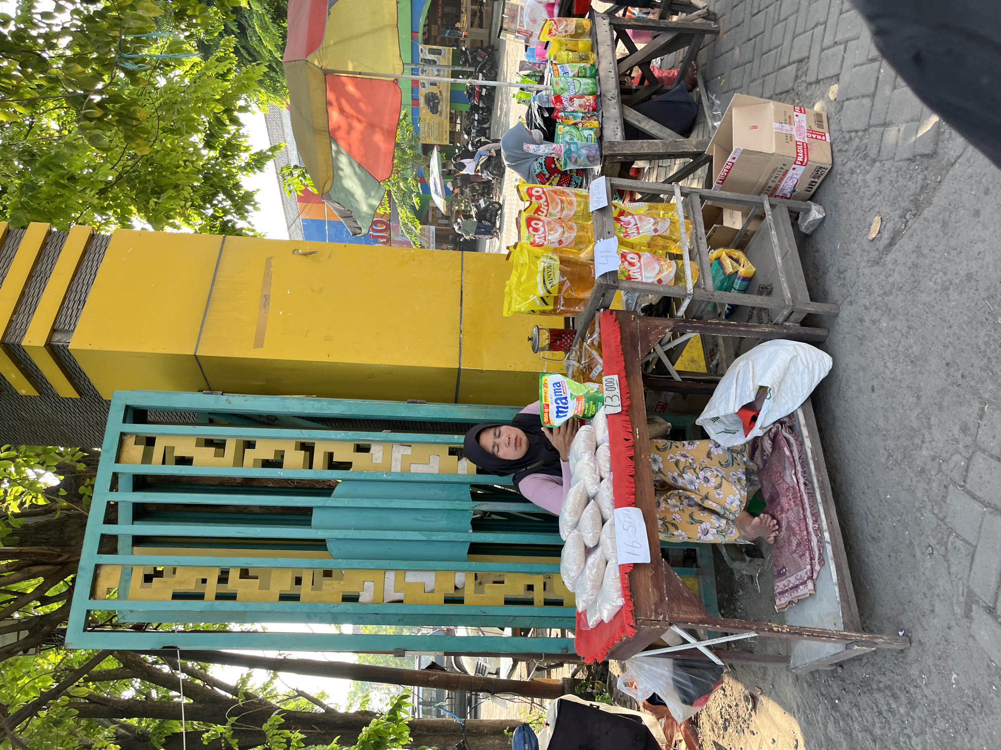 This image shows a street vendor in Juwet Kenongo, Indonesia, selling goods from a wooden cart. The vendor, a woman wearing a black hijab and a pink top, sits behind a red-cloth-covered table laden with white-bagged merchandise, possibly rice. A sign on the table reads "16.500". Adjacent to her cart, a yellow pillar and a turquoise and yellow gate stand prominently. On a tiered shelf to her right, various yellow and red packaged goods, including cooking oil, are displayed, along with a sign indicating "13.000". A colorful umbrella shades some of the merchandise. In the background, other vendors, motorbikes, and people are visible, suggesting a bustling market environment. The ground is paved with grey cobblestones, and trees with green foliage provide shade overhead. The scene depicts a typical street market setting in a populated urban area.