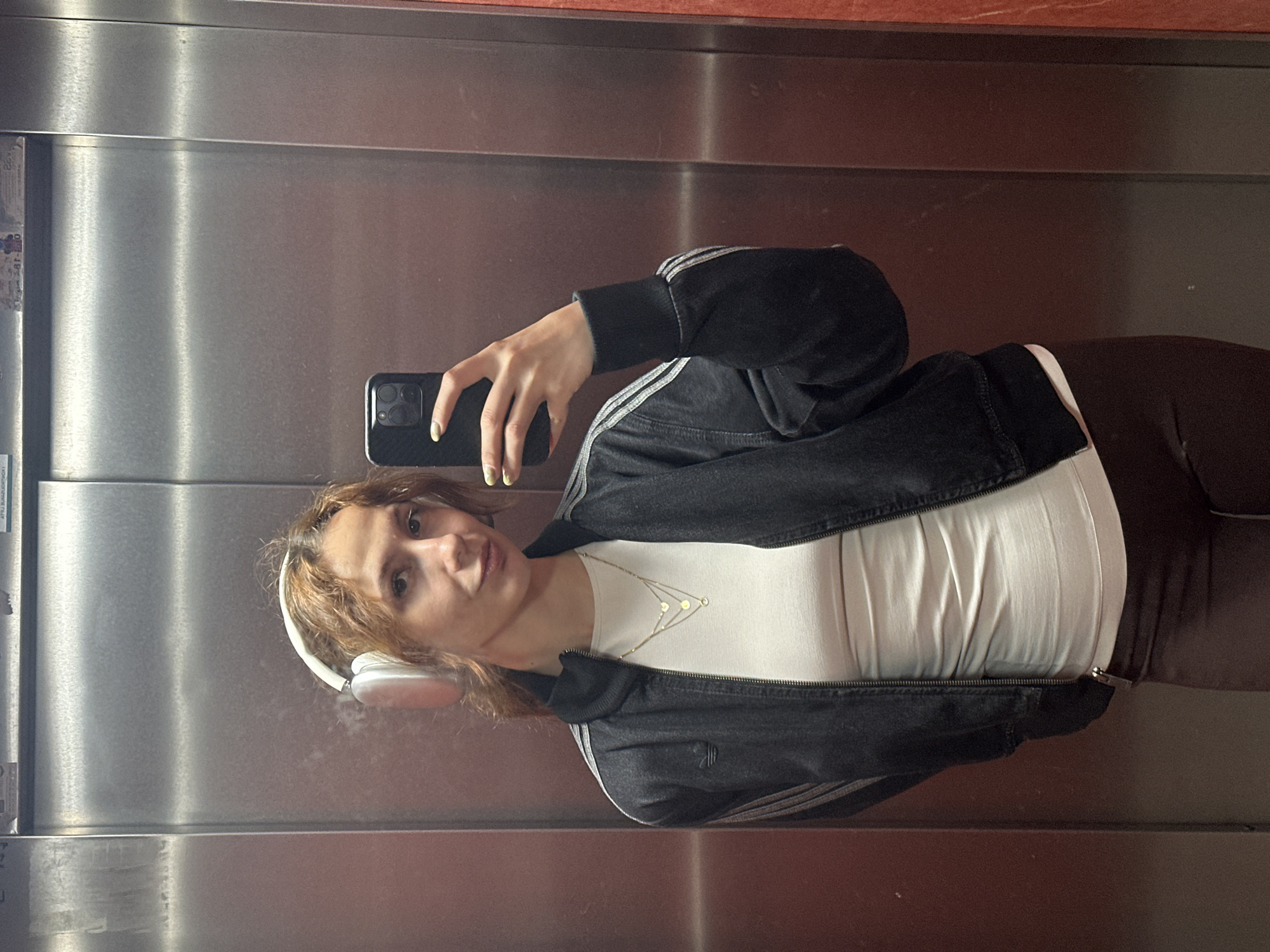 A young woman is taking a selfie in an elevator. She is wearing headphones, a black Adidas track jacket, a white top, and brown leggings. She is holding a phone in her right hand. The elevator has stainless steel walls. The image was taken in Novi Sad, Serbia.