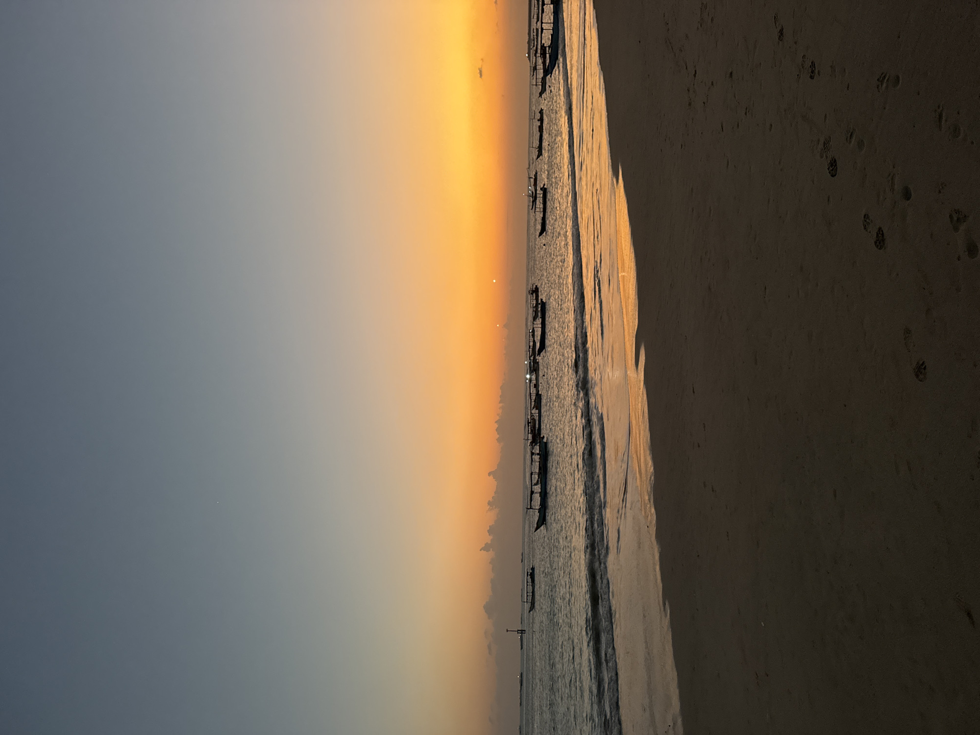 A wide shot captures a beach at dusk in Kuta, Indonesia. The sky transitions from a deep blue at the top to a vibrant orange and yellow gradient near the horizon, indicative of a sunset. A few scattered clouds are visible, particularly lower down. The sea is calm with gentle waves lapping the shore. Several traditional Indonesian fishing boats are anchored or adrift on the water, silhouetted against the colorful sky. The foreground is a sandy beach, with footprints visible in the damp sand near the water's edge. The overall atmosphere is tranquil and serene.