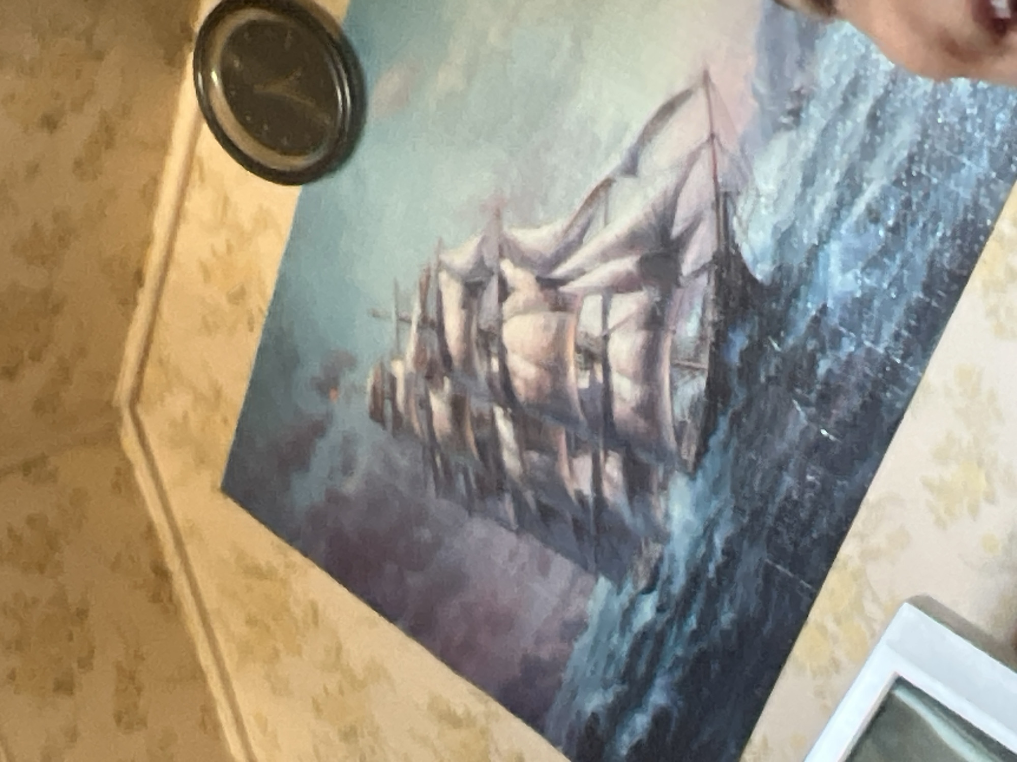 A painting of a tall ship with full sails is depicted in rough seas. The ship is angled to the right, with waves crashing around it. The sky is dark and stormy, with dramatic clouds. To the upper right of the painting, a black circular object with a dark face and light-colored hands is visible; it appears to be a clock. The painting is placed on a surface that has a textured, patterned wallpaper in a light beige or cream color. A portion of a person's face is visible in the lower right corner, with their mouth slightly open. The scene is lit by an unseen light source, casting shadows and highlights on the objects and the painting. The overall impression is one of a maritime scene, possibly a historical painting. The location in Krasnoslobodsk, Russia, is not evident from the visual information provided in the image.