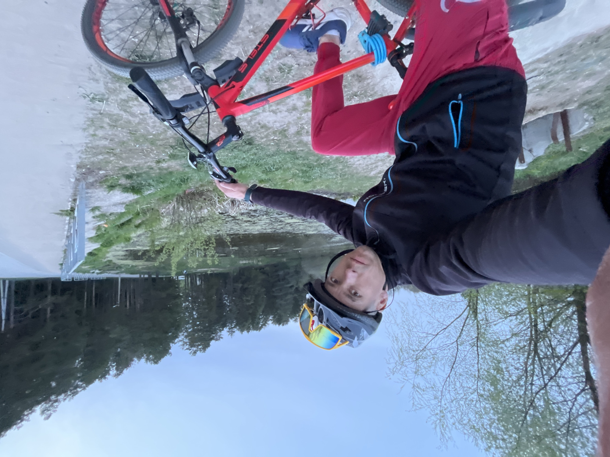 A man is on a red mountain bike near a body of water, likely a lake or wide river, in the city of Chaikovsky, Russia. The man is wearing a helmet and sunglasses and is dressed in a black jacket and red pants. He appears to be taking a selfie. The bike is resting with one pedal anchored by a blue object. The background features trees and possibly buildings along the shore, indicating a park or recreational area. The image is rotated 180 degrees.