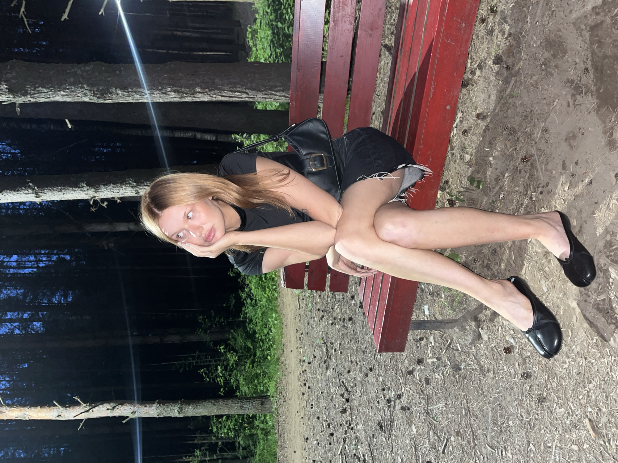 A young woman with blonde hair is sitting on a red park bench in a forest at night. She is wearing a black t-shirt and a black denim skirt. She has on black split-toe shoes. A black handbag with a gold buckle rests on the bench next to her. The ground around the bench is covered in pinecones and dirt. Trees with thin trunks are visible in the background, with faint lights illuminating the forest. The woman is looking up and to her left, with her hand resting on her cheek. The image was taken in the city of Chaikovsky, Russia.