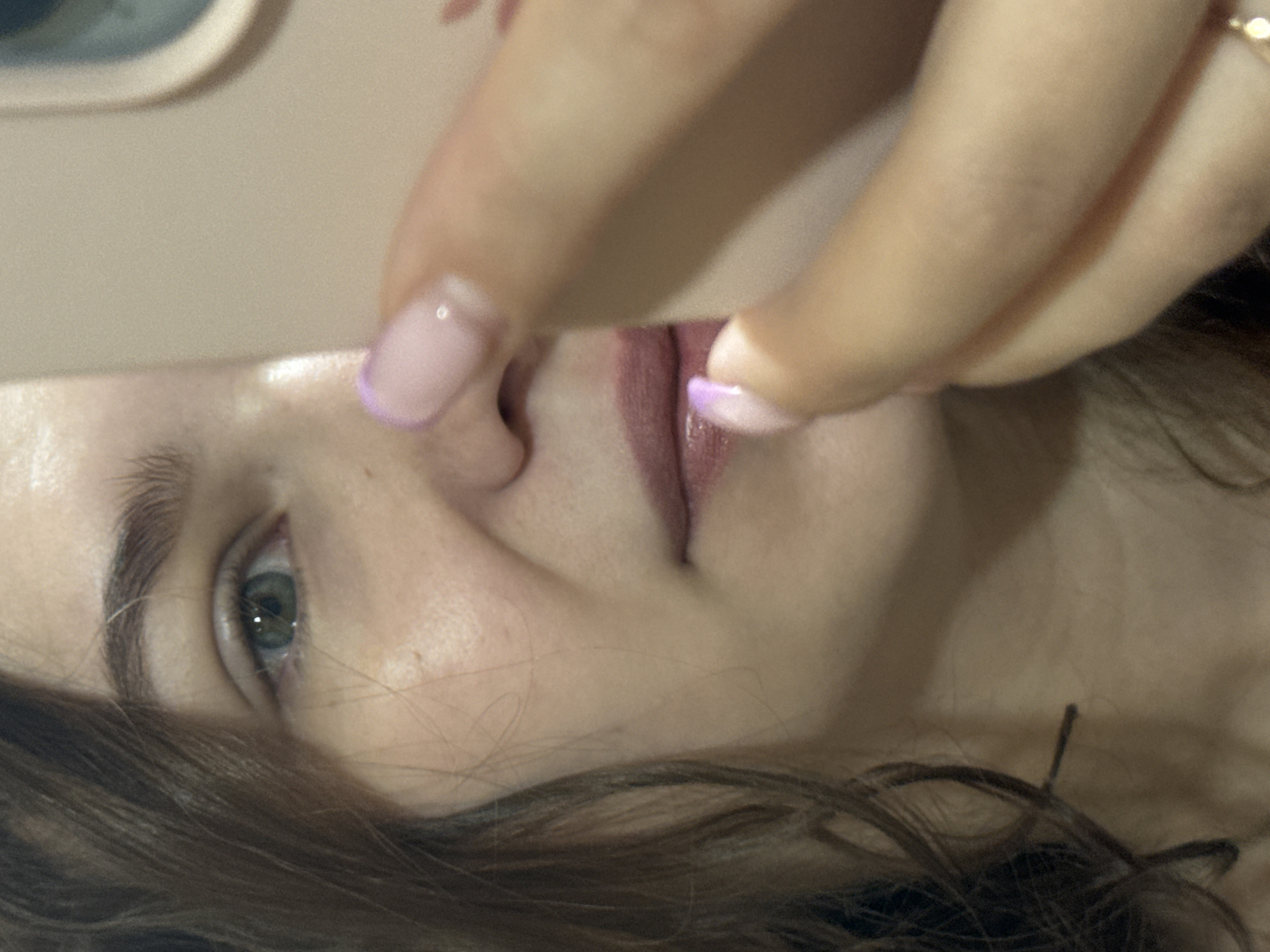 A close-up, off-kilter shot depicts a person's face, partially obscured by fingers with light pink and purple-tipped nails. The person has fair skin, light eyebrows, and a visible eye with light green irises. Some brown hair is visible near the bottom of the frame. A tan-colored, flat object (possibly a phone) covers the upper left portion of the face. The person's lips are visible, appearing to be wearing lip gloss. The image contains no discernible information that places it in Nizhnyaya Gar', Russia, or any other specific location.