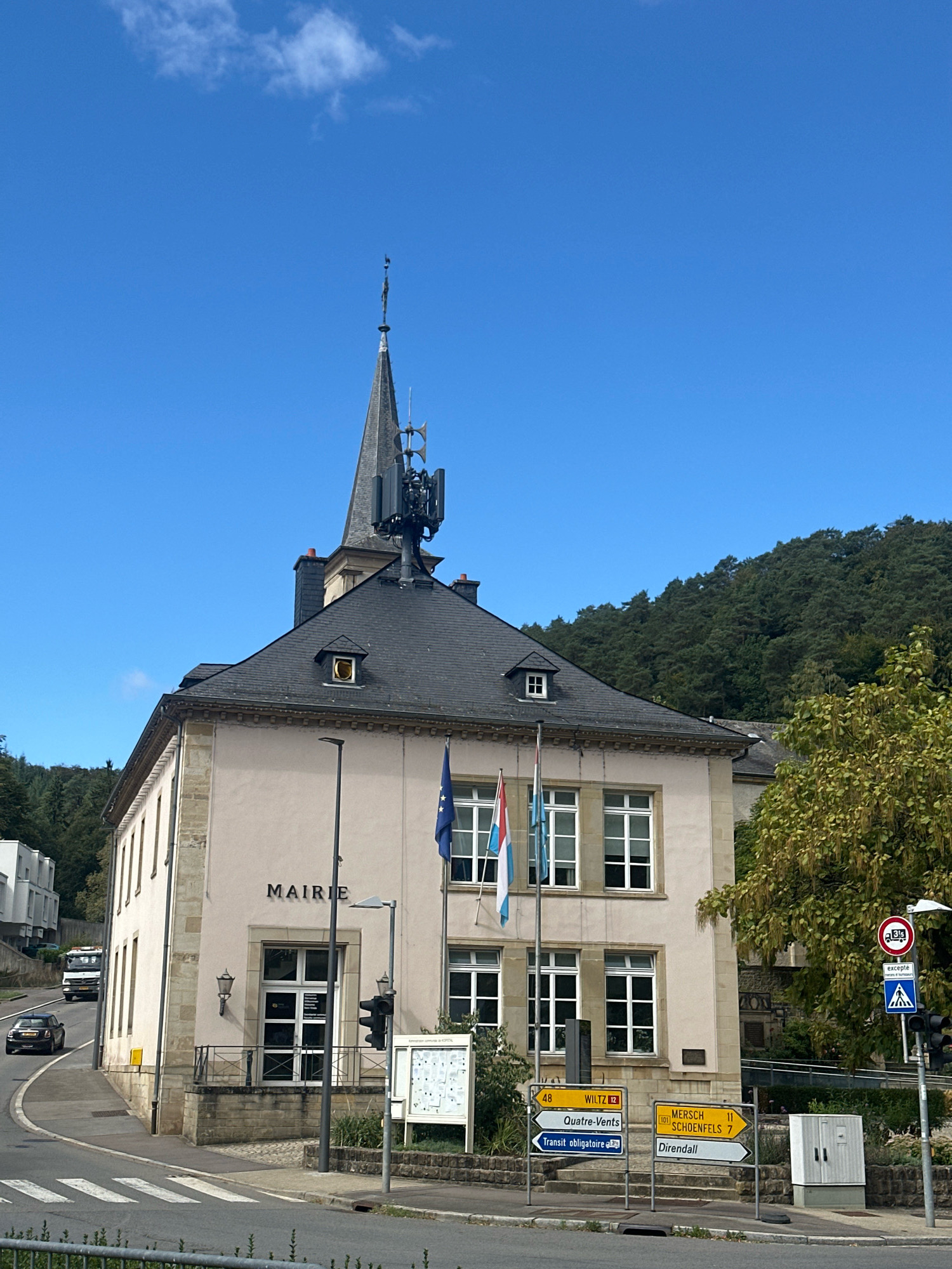 In the city of Bridel, Luxembourg, a two-story building with a light-colored facade displays the word "MAIRIE" in large black letters above its entrance. The building features numerous multi-paned windows and a dark-tiled roof, topped by a tall, dark spire that has a modern antenna array attached. Flags are displayed on the building's facade. In front of the building, a sidewalk with a railing leads to the entrance. To the right of the building, various road signs indicate directions and distances to towns such as Wiltz, Quatre-Vents, Mersch, Schoenfels, and Direndall, along with a pedestrian crossing sign and a traffic signal. A utility box is situated near the signs. The scene is set against a backdrop of a lush, tree-covered hillside under a clear blue sky with a few wispy clouds. A road curves in front of the building, with a pedestrian crossing marked. A dark-colored car is visible on the road, and a white truck is further up the road on the left. The overall impression is one of a well-maintained civic building in a semi-rural setting.