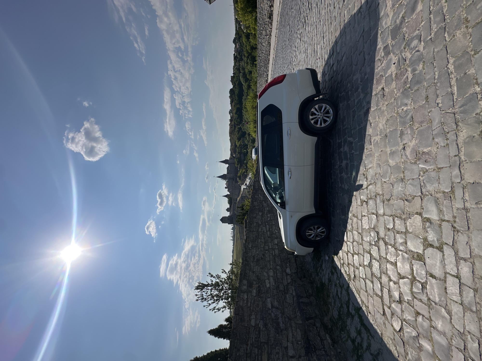 A white SUV is parked on a cobblestone street in Kamianets-Podilskyi, Ukraine. The vehicle is positioned in the foreground on the right side of the image, facing left. Behind the SUV, a stone wall runs along the left side of the street. In the background, a historical castle complex with multiple towers and spires is visible on a hill, surrounded by green trees. The sky is blue with scattered white clouds and a bright sun with lens flare is visible in the upper left corner. The cobblestone street shows signs of wear and is bathed in sunlight, casting shadows. The overall scene suggests a sunny day in a historic European city.
