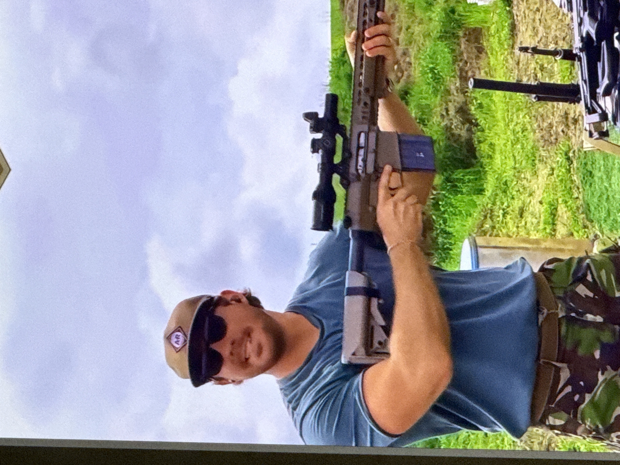 In an outdoor setting under a cloudy sky, a light-skinned man wearing a blue t-shirt, camouflage pants, sunglasses, and a brown baseball cap with an "AR" logo is holding a rifle with a scope. He is smiling and looking to his left. The rifle has a magazine inserted. In the foreground, other firearms are visible. The background consists of green grass and a dirt embankment. There are no discernible landmarks or features to indicate the specific location in the city of Cahul, Moldova.