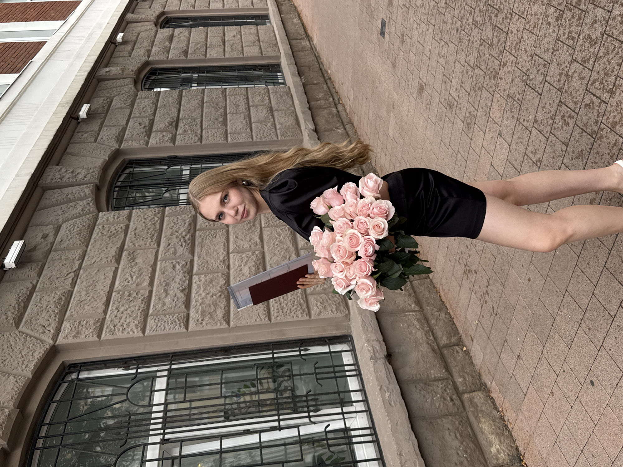 A young woman with long blonde hair is standing on a sidewalk next to a stone building. She is wearing a short black dress and holding a bouquet of pink roses in her left arm and a clear plastic case with a red document inside in her right hand. The building has arched windows with black metal grates. The sidewalk is made of brick pavers. The scene appears to be in a city, possibly a university campus or a historic district. There is no explicit information in the image to confirm the location in Perm, Russia.