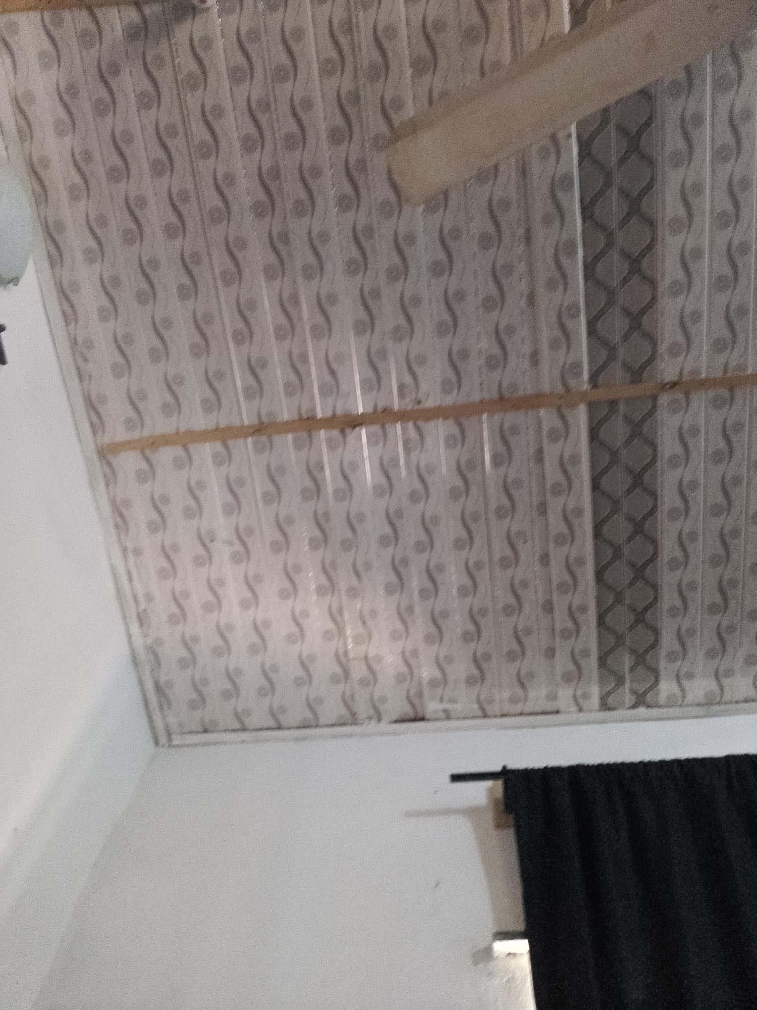 The image shows a low-angle view of a ceiling with a patterned design of wavy lines and circular shapes. There are two horizontal wooden beams crossing the ceiling. In the lower right portion of the image, a black curtain is visible hanging from a rod. The walls appear to be white. The scene is indoors. The location of Jauro Yinu, Nigeria is not determinable from the visual information provided in the image.