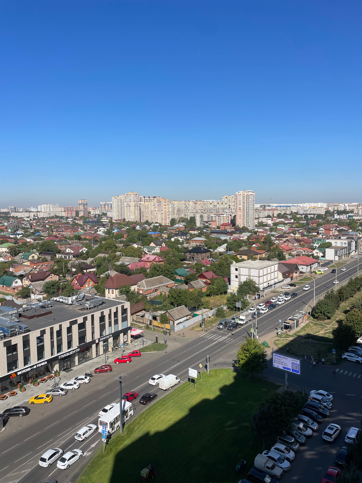 The image provides an aerial view of a mixed urban and suburban landscape under a clear blue sky in Krasnodar, Russia. The foreground features a modern, multi-story building with a striped facade and storefronts, including one labeled "ГЛАЗЦЕНТР" and another "ВКУСНО-И ТОЧКА". Several cars are parked along the sidewalk in front of this building and on the adjacent street.
The street itself is a multi-lane road with moderate traffic, including passenger cars, a white van, and a white bus. Cars are also seen parked on the opposite side of the street and on a side road. A pedestrian crossing is visible at an intersection.
Beyond the immediate street level, the scene transitions to a dense neighborhood of single-family homes with red, brown, and green roofs, interspersed with numerous trees. In the background, the skyline is dominated by numerous high-rise apartment buildings, indicative of a more densely populated urban area. The overall impression is a snapshot of a developing city with a mix of residential types and commercial activity.