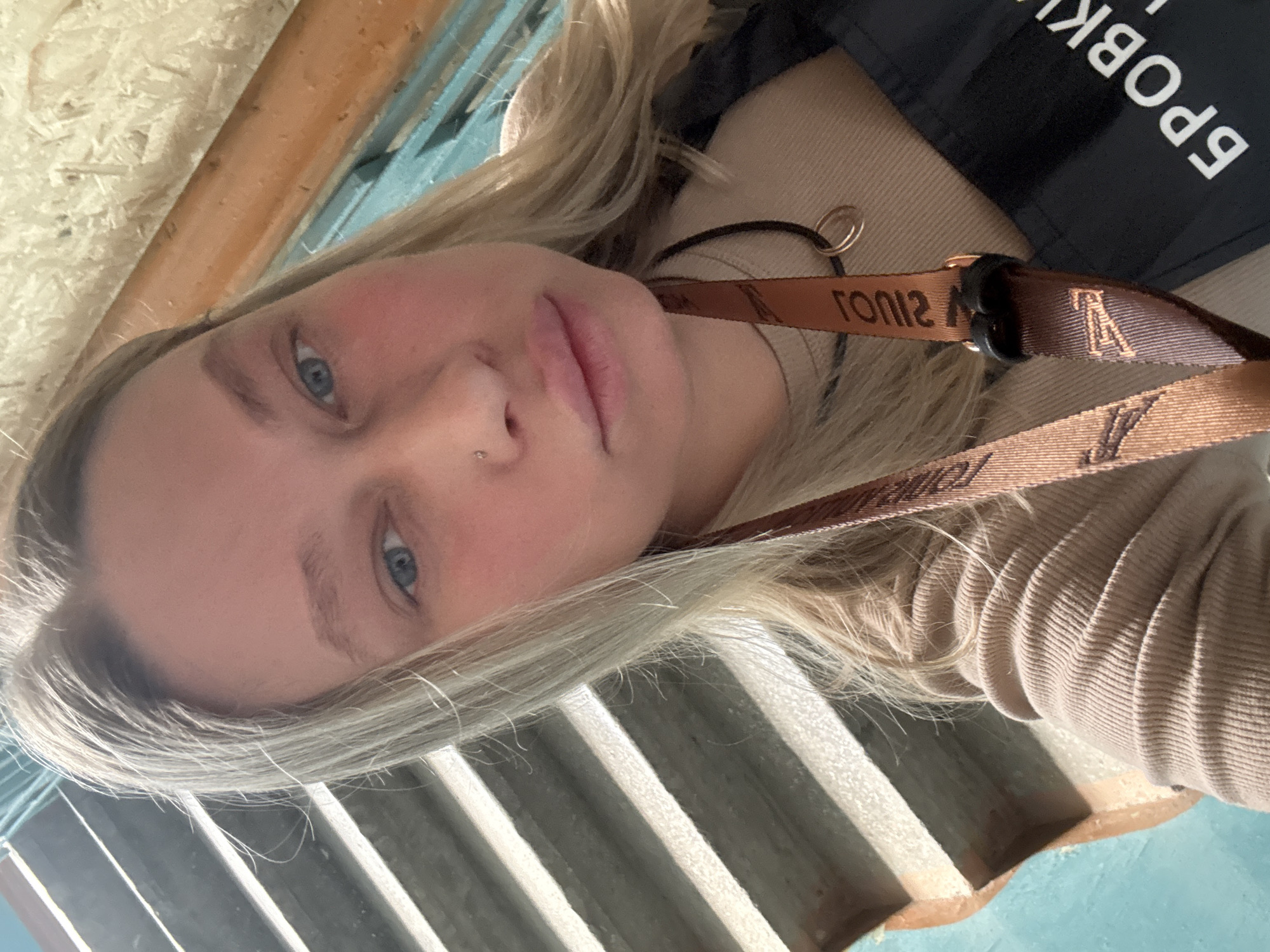 The image is a tilted, close-up selfie of a young woman with long, light blonde hair and blue eyes. She has fair skin, light makeup, and a small silver piercing on her left nostril. Her lips are slightly parted, and her expression appears neutral and calm, with her gaze directed towards the viewer.
She is wearing a light beige, ribbed top or sweater, visible on her right side. Around her neck, a brown strap with the "Louis Vuitton" monogram and the text "LOUIS" is visible, suggesting it's part of a branded accessory like a lanyard or bag strap. Over her right shoulder, a black strap or bag is present, featuring white Cyrillic text that clearly spells out "БРОВКИ" (BROVKI), which translates to "eyebrows" and is often associated with beauty services.
The background is fragmented and suggests an indoor setting. In the upper left portion, a textured surface resembling light-colored wooden particle board or oriented strand board (OSB) is visible, along with a smoother light brown wooden element and a bright turquoise or light blue surface. In the lower right, a series of light-colored, ridged or slatted panels, possibly part of a wall or heating vent, can be seen. The overall lighting suggests a well-lit indoor environment, likely during daytime. The location context for this image is Perm, Russia.