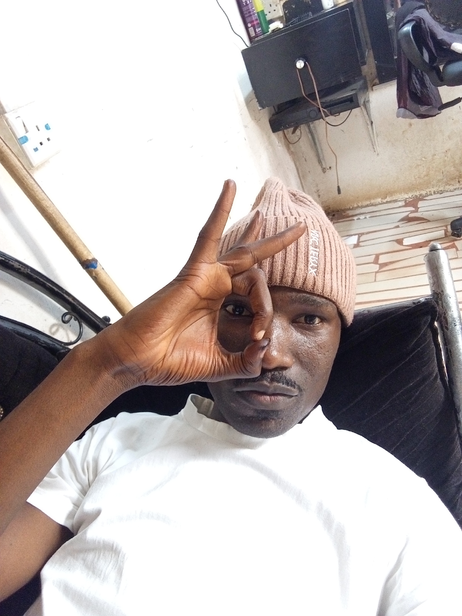 A man is sitting in a dark room in Gwong, Nigeria, making an “OK” sign with his hand over his right eye. He is wearing a tan knit beanie and a white t-shirt. Behind him is a black padded object with a metal frame. In the background, there is a wall with a power outlet and a shelf with a television. The walls are somewhat discolored with marks. A wooden rod leans against the wall.
