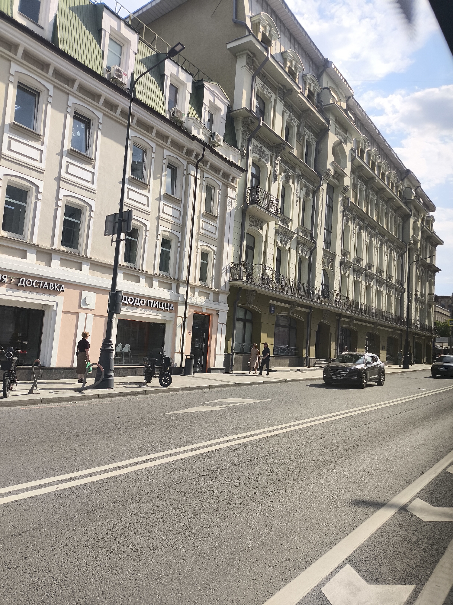 The image depicts a street scene in Moscow, Russia. The buildings along the street are multi-story, with detailed architectural designs. One building houses a "Dodo Pizza" restaurant with a pink facade and delivery service. Cars are visible on the multi-lane street, including a black Hyundai Tucson with a license plate ending in "E405PE 190". Several pedestrians are walking on the sidewalk. Street lights and other infrastructure elements are also present.
