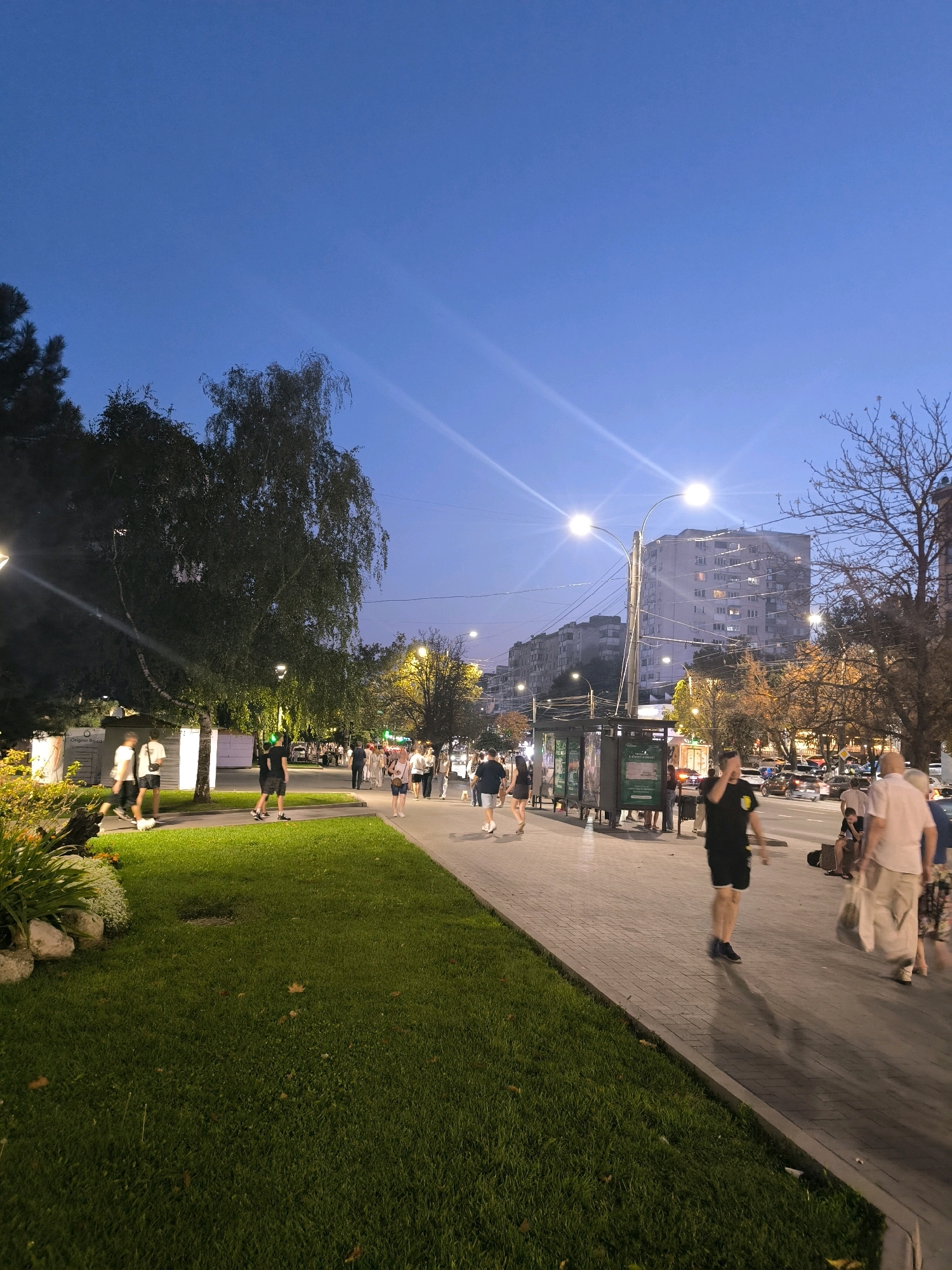 A street scene in Chișinău, Moldova, is captured at dusk. A well-lit sidewalk runs along a grassy median strip, with a road for vehicular traffic on the right. Several streetlights illuminate the area. On the sidewalk, various individuals are walking, some alone and others in groups. A man in a black t-shirt and shorts is walking towards the viewer, casting a long shadow. A woman in a light-colored top and patterned skirt is walking away from the viewer. Further down the sidewalk, more people are visible, and a bus stop shelter is present. Across the street, buildings, trees, and cars are visible. The sky is a deep blue, indicating the late hour.